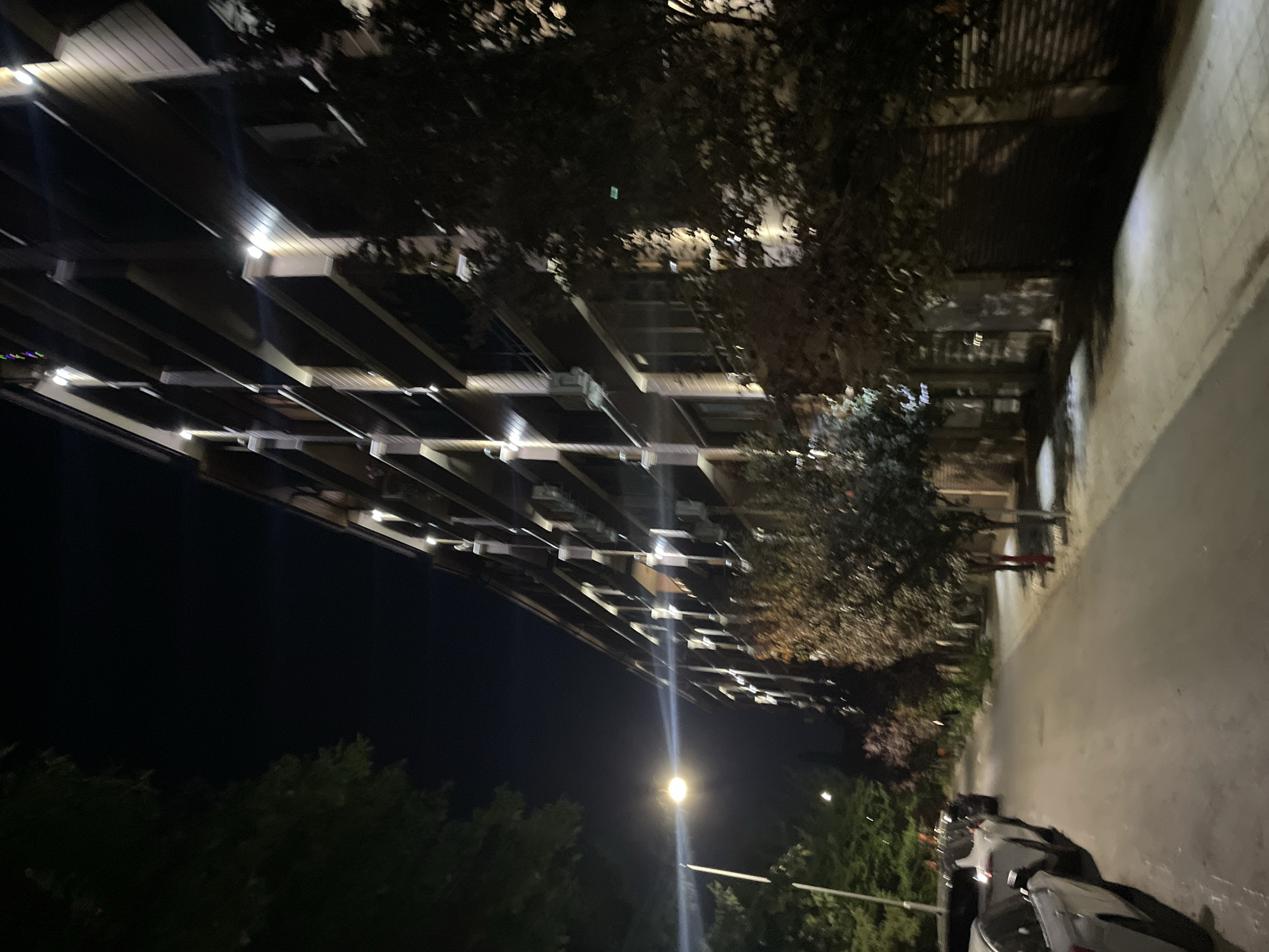 The image depicts a nighttime street scene in Burgas, Bulgaria. A multi-story apartment building with illuminated balconies runs along the right side of the frame. The building appears to be modern, with vertical white columns supporting each balcony. Trees and foliage line the street, casting shadows on the pavement. Several parked cars are visible on the left side of the street, facing away from the camera. A street lamp illuminates a portion of the road and sidewalk. The overall atmosphere is dark and quiet, suggesting a residential area at night.