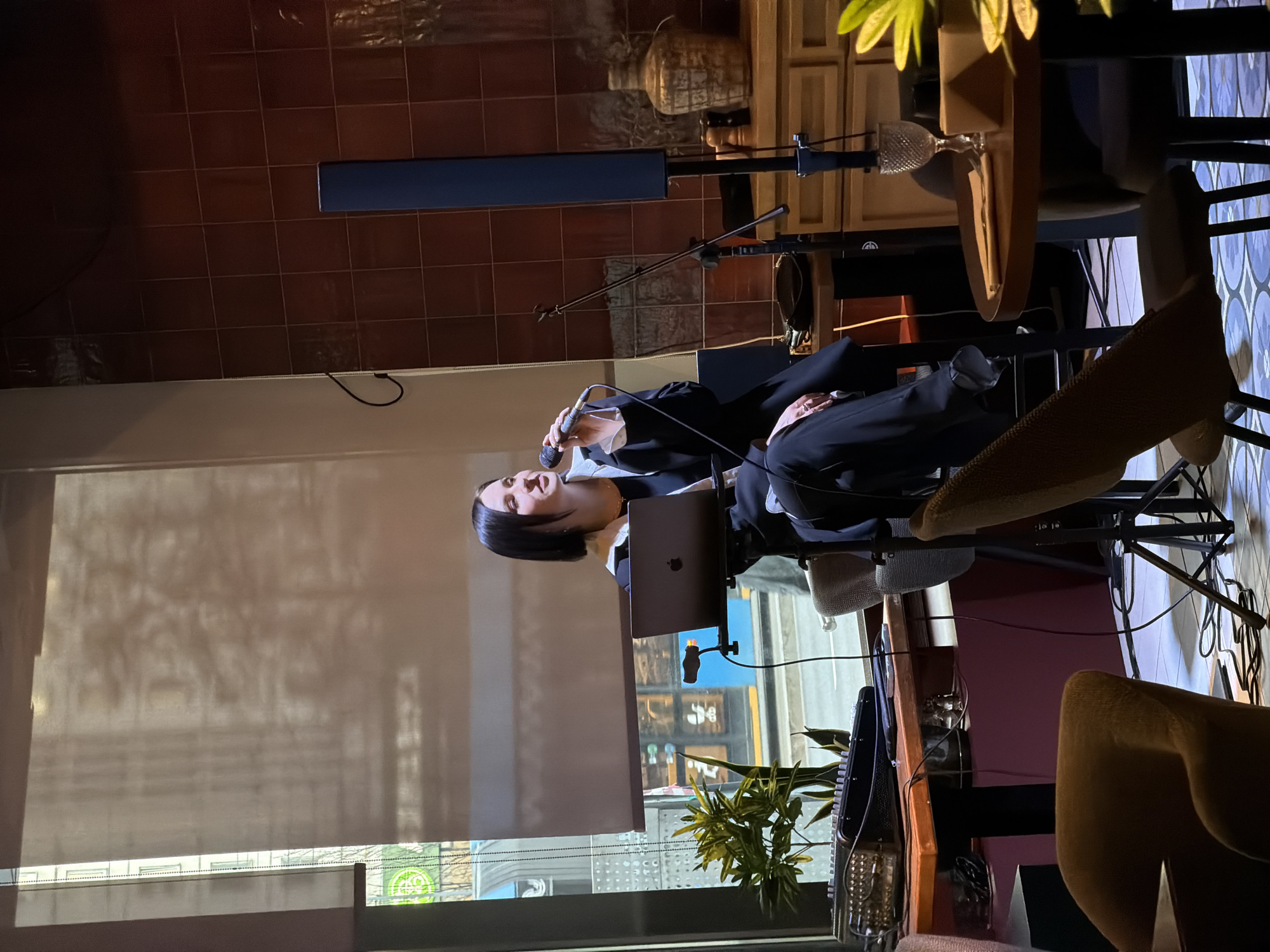A woman in a dark blazer and white shirt is giving a presentation or performance. She is holding a microphone and standing behind a laptop computer, which is positioned near a window. The window provides a view of a city street, which may be in Moscow, Russia, given the architecture and context. The street scene includes shops and other buildings. The interior setting appears to be a restaurant or performance space, with dark wood paneling and tile flooring. There's a microphone stand and seating nearby, suggesting an area for live music or presentations.