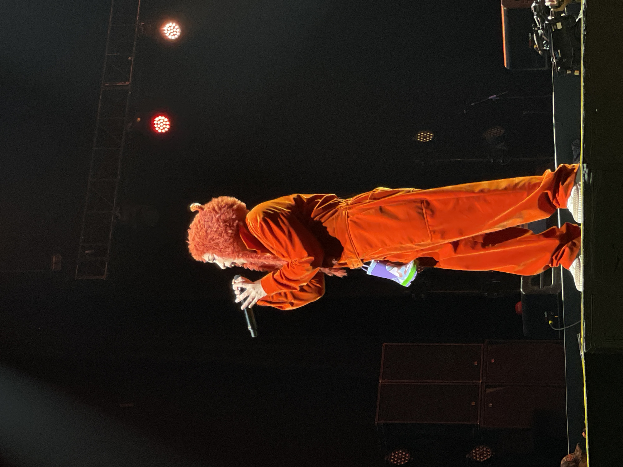 A person wearing an orange jumpsuit and a fuzzy orange head covering is holding a microphone and singing on stage in front of an audience. There are stage lights and sound equipment visible in the background. The location is Salembaran, Indonesia.