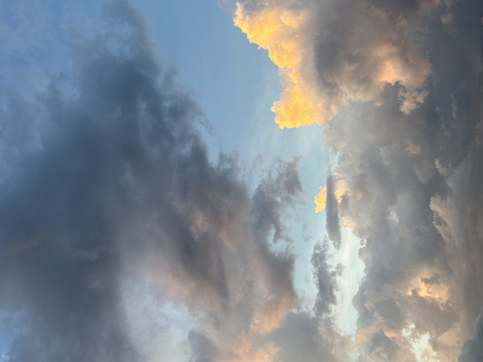 The image depicts a dramatic sky filled with a mix of dark and light clouds, illuminated by what appears to be the setting or rising sun. The clouds range from deep grays and blues to soft pinks and vibrant yellows, creating a sense of depth and movement. The sky is a clear blue in some areas, contrasting with the denser cloud formations. This celestial scene is likely captured from an elevated viewpoint, possibly a building or natural landmark, offering a panoramic view of the sky over the city of Đà Nẵng, Vietnam. The absence of any visible landmarks or specific city features prevents definitive identification of the precise location within Đà Nẵng. The focus is entirely on the atmospheric conditions.