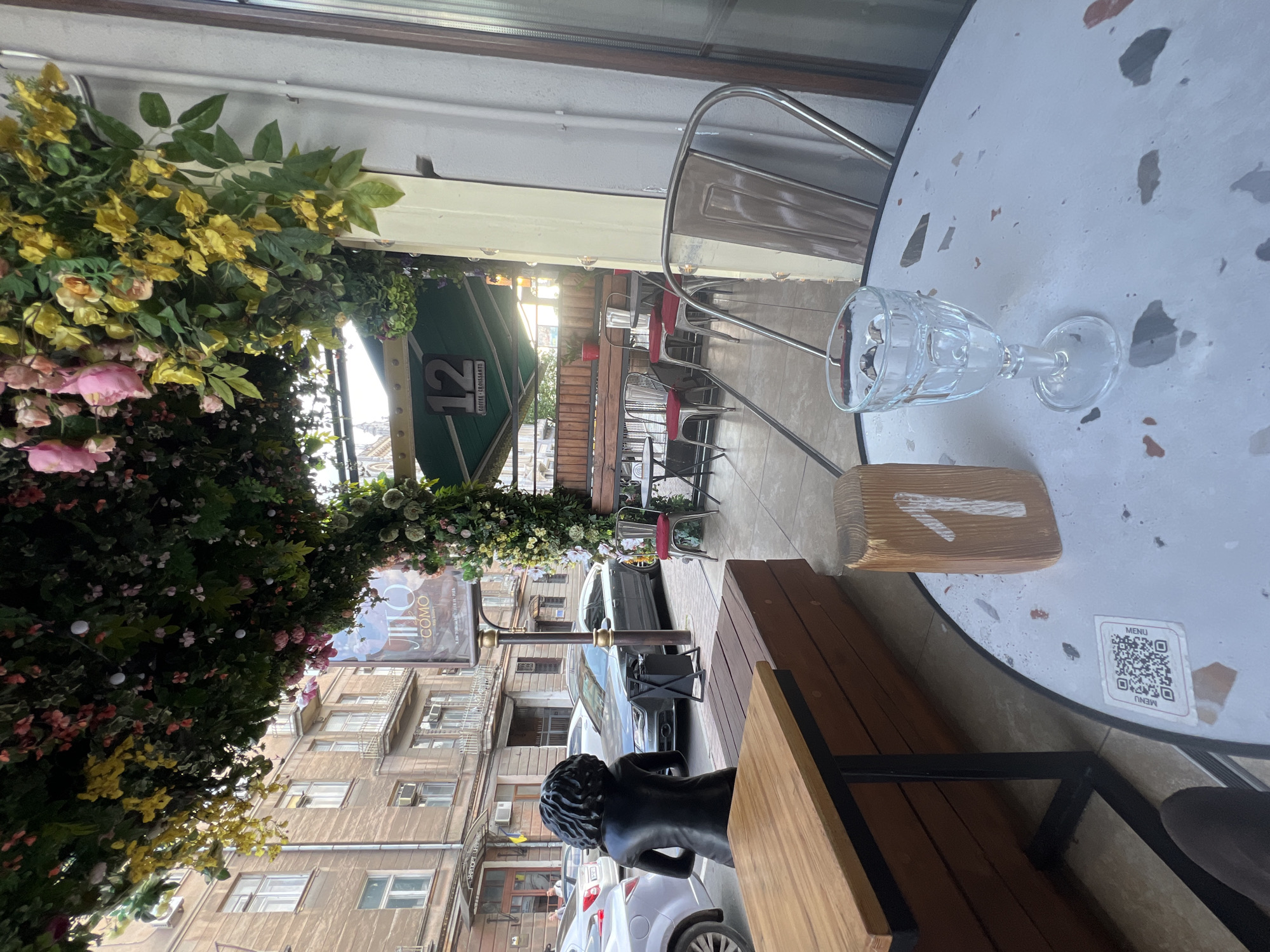 The image depicts an outdoor seating area of a cafe in Odesa, Ukraine, as indicated by a banner displaying "12 Coffee Croissants" and a street view with European-style architecture. The foreground features a white speckled table with a clear glass of water and a wooden table number stand with a white '1' painted on it. A QR code labeled "MENU" is visible on the table. To the left, a wooden bench is situated next to a wooden table with a black metal frame. In the background, a dark-colored statue of a person with curly hair faces away from the camera. Beyond the seating area, cars are parked on the street, and a building with multiple windows and balconies lines the opposite side of the street. An awning covered in artificial flowers and greenery shades the cafe's seating area.