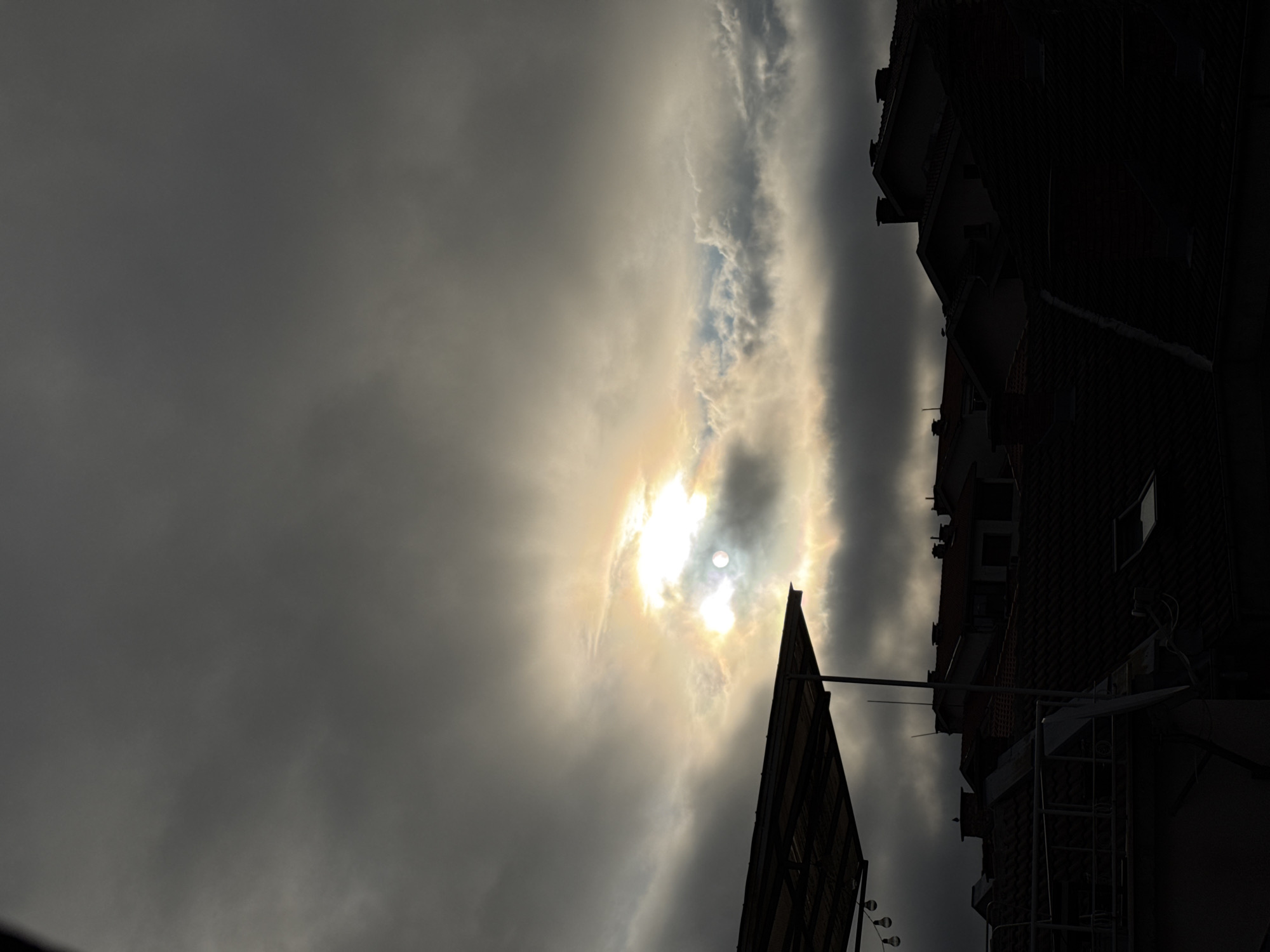 The image shows a view of rooftops in Pleven, Bulgaria, under a dramatic, overcast sky. The sun is partially obscured by dark clouds, creating a bright, diffused light that illuminates a patch of sky with a yellow and white glow. Some lighter, wispy clouds are visible. In the foreground, a dark, silhouetted structure, possibly a canopy or awning, juts in from the left side of the frame, with a series of light bulbs hanging from it. Below the canopy, a row of tiled rooftops of houses and buildings is visible, mostly in shadow. Chimneys and skylights are discernible on the roofs. The overall impression is of a moody, possibly late afternoon or early morning scene in a residential area of the city. No people are present in the image.