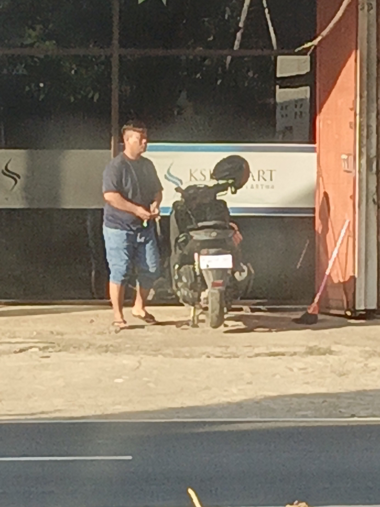 In the city of Srono, Indonesia, a man stands next to a parked scooter outside a building. The man is wearing a dark t-shirt and light blue shorts. He is holding a green object in his right hand. The scooter is black and has a license plate. A pink and white mop is leaning against the wall of the building. The building has a red facade on the right side and a large sign in the background with text that appears to read "KSK ART". The scene is illuminated by sunlight, casting shadows on the ground. The road is visible in the foreground.
