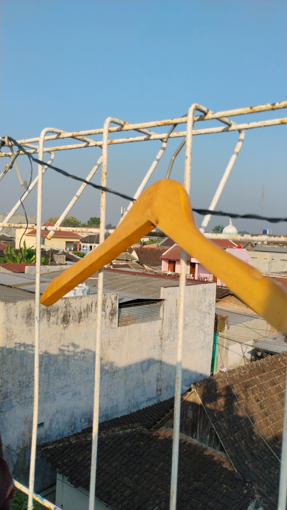 A wooden clothes hanger is suspended from a white, rusty metal railing against a clear blue sky. The railing is part of a drying rack or balcony enclosure. In the background, a densely packed urban landscape of residential buildings is visible. The rooftops are predominantly tiled, with some buildings painted in light colors, including a notable pink structure with brown trim. Vegetation, including green trees, is interspersed among the buildings. A white dome structure, possibly a mosque, is visible in the distance, along with a telecommunication tower. The scene appears to be set in a village or town, likely during daylight with the sun casting shadows. The image was taken in Gumpang, Indonesia.