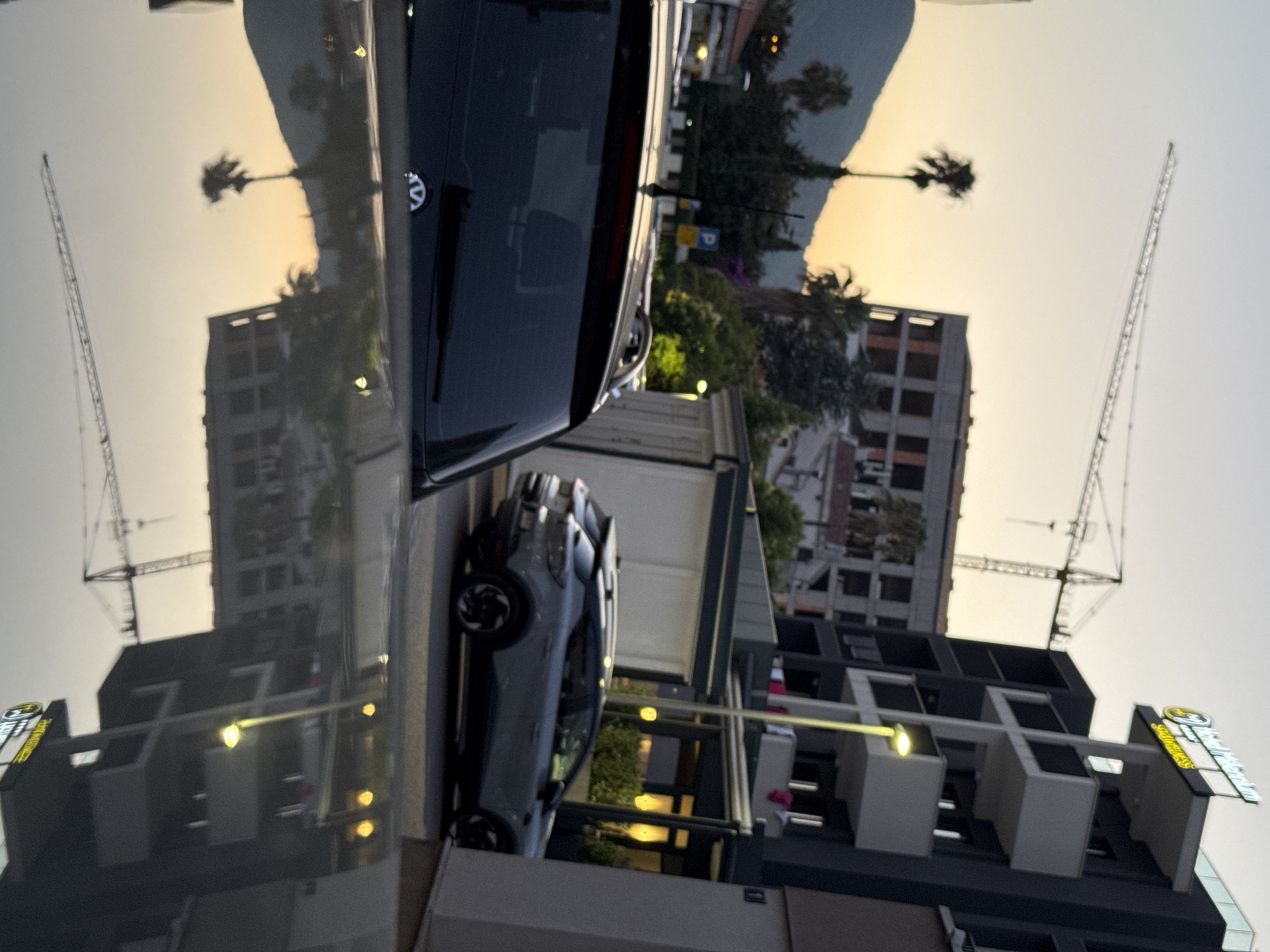 The image depicts a street scene in Budva, Montenegro, during twilight. A modern, dark-colored building with multiple balconies and a prominent "Hotel Millennium" sign is visible on the right. A construction crane is present in the background, suggesting ongoing development. Several cars are parked on the street, including a grey hatchback in the foreground. The reflection of the scene on the car's hood shows palm trees, the building, and the crane. The overall atmosphere is calm, with the warm glow of streetlights and the setting sun creating a serene ambiance.