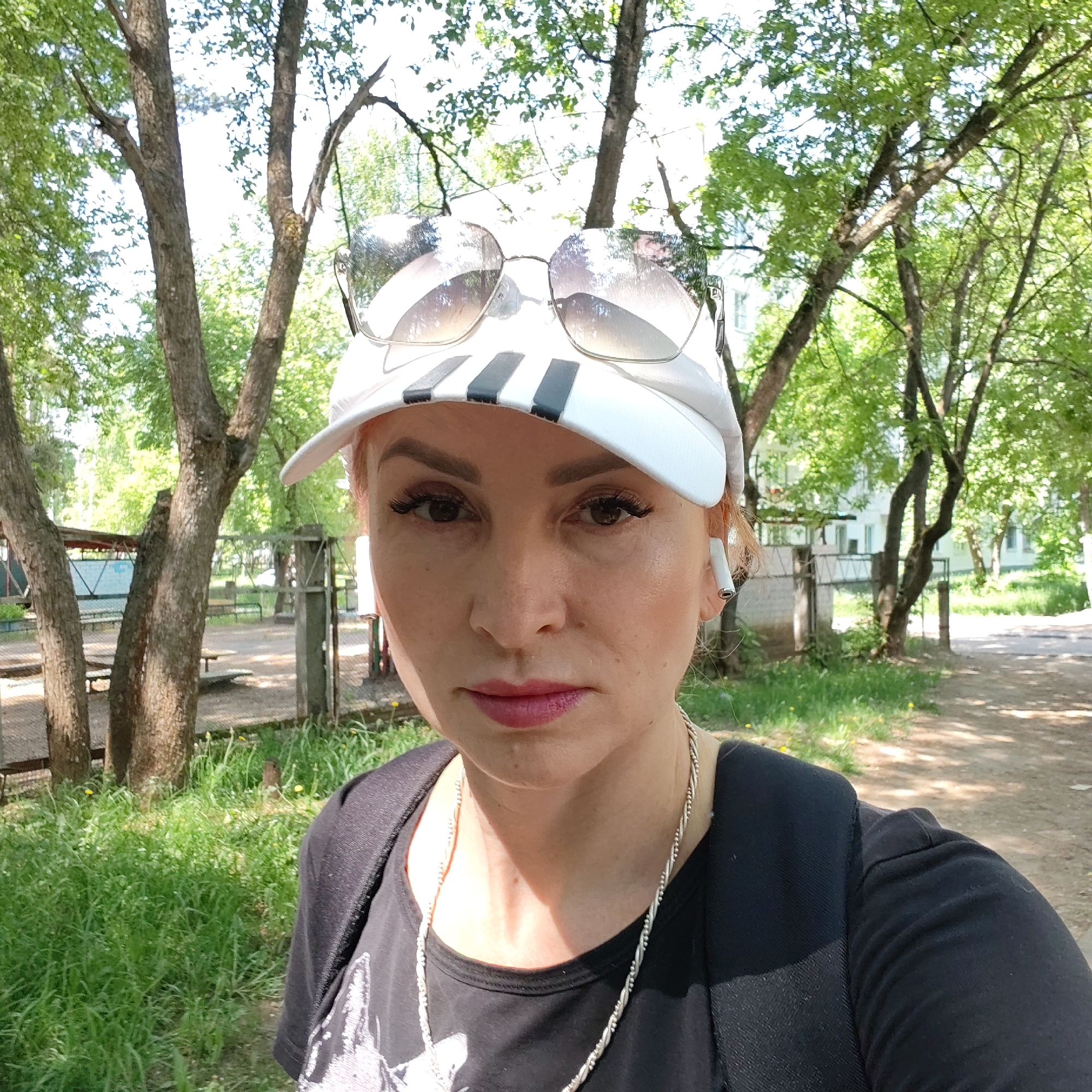 A selfie shows a middle-aged woman with reddish-blonde hair wearing a white baseball cap with black stripes, sunglasses perched on top of the cap, a black t-shirt, and a silver necklace. She has fair skin, dark eyelashes, and pink lipstick. She has a white wireless earbud in her right ear. The background shows a park-like setting with trees, grass, and a dirt path. A fenced area with picnic tables is visible behind the woman. The location is outdoors and appears to be in a residential area in Chaikovsky, Russia, as indicated by the style of the buildings and the overall appearance of the environment. The woman is the primary subject of the image, with the park setting acting as the background.