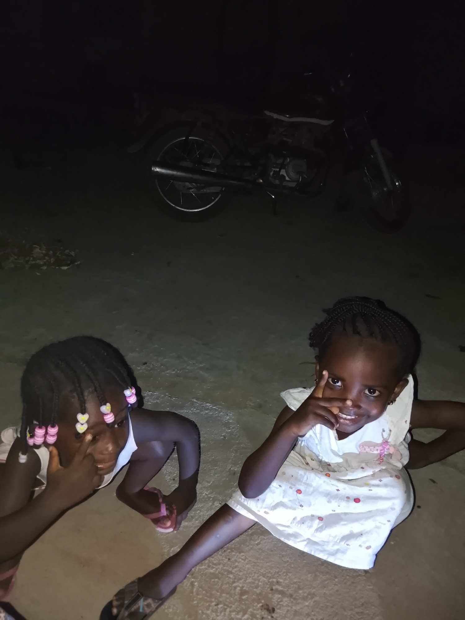 Two young girls are squatting on a concrete surface in Akwanga, Nigeria. In the background is a parked motorcycle. The girl on the right is wearing a light-colored patterned dress, and she has a finger pointed toward her face. The girl on the left is wearing a light-colored shirt and has colorful beads in her braided hair. She also has a finger near her face. They are both looking at the camera. It is night, and the area is dimly lit.