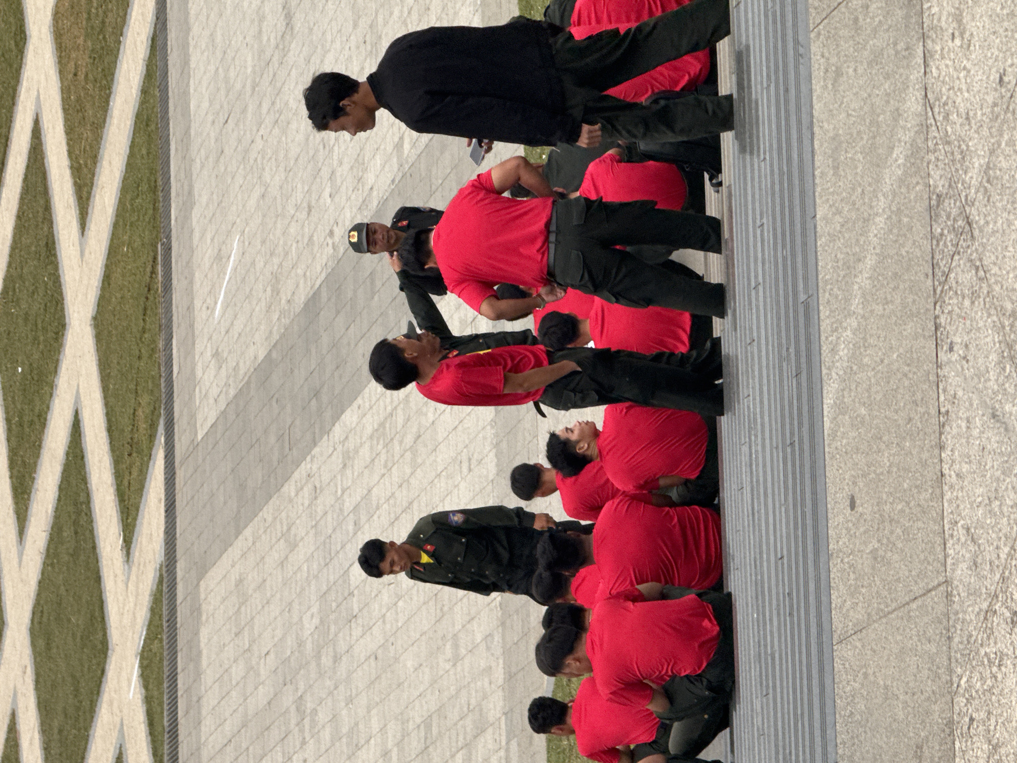 The image shows a group of young men in uniform, some wearing red shirts and black pants, others in a dark green military uniform. They appear to be in formation or participating in a drill. The setting is outdoors, on a paved area with a section of grass and a concrete border. The paved area is made up of large, light-colored tiles. In the background, there are diagonal white lines on the grass, forming diamond shapes. Some of the men are kneeling, while others are standing or in a staggered position. One individual in the dark green uniform appears to be leading or instructing. The lighting suggests it is daytime, and the shadows cast by the people indicate the sun is at a moderate angle. The overall atmosphere seems to be one of discipline and training. There is no visible text in the image.