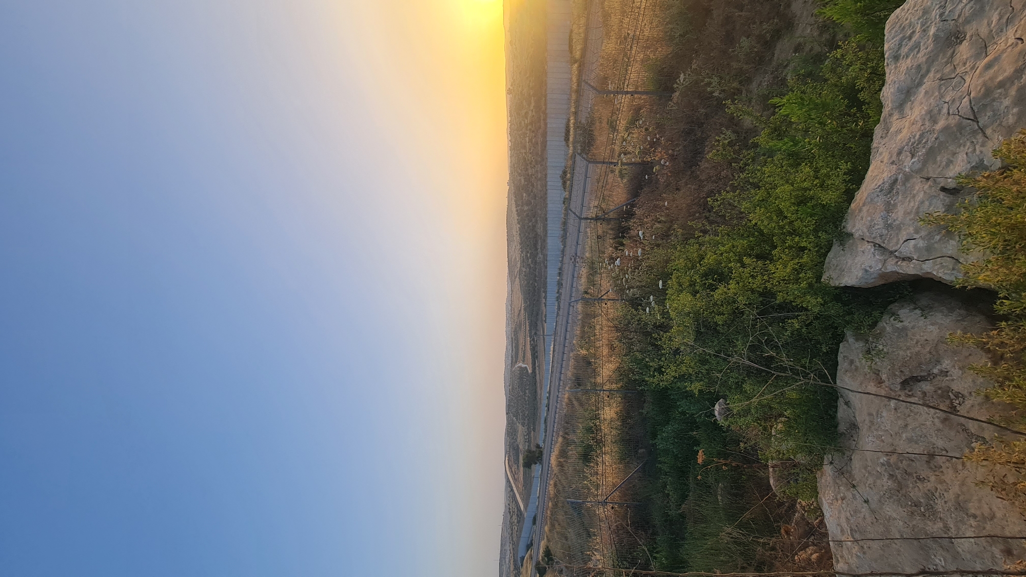 The image shows a sunset landscape, possibly in the Lachish Regional Council, Israel. On the right side of the image are light-colored rocks and green vegetation. To the left of this, there appears to be a long fence and some sort of structure, potentially a wall or barrier, that stretches into the distance. The sky is blue transitioning to a yellow-orange glow at the top where the sun is positioned. The landscape consists of dry vegetation and fields.