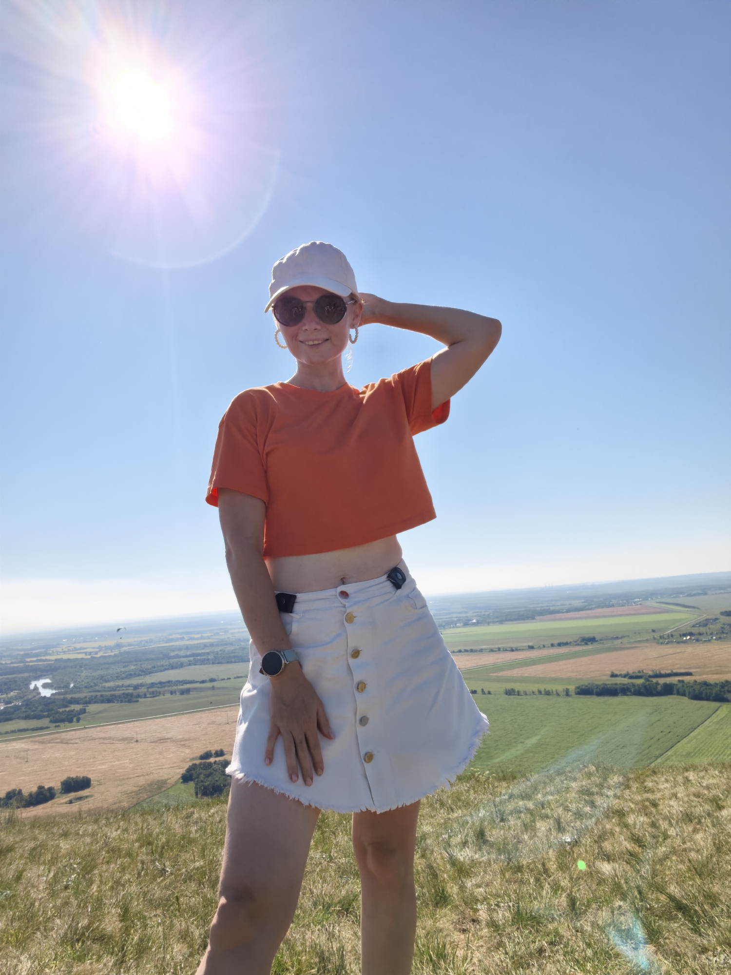 A woman is standing on a grassy hill overlooking a rural landscape under a bright, sunny sky. She is wearing an orange crop top, a white denim skirt, a white baseball cap, and sunglasses. Her left arm is raised and resting on her head. A smartwatch is visible on her left wrist. The background shows fields, a river, and a distant town. The image appears to be taken in the vicinity of Sterlitamak, Russia, possibly from a viewpoint like the Shikhany mountains.