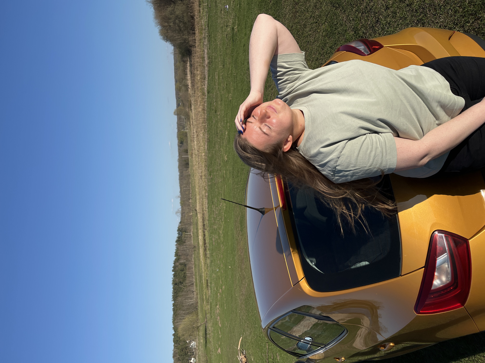 A woman is lying on the hood of a golden car, with her head resting on her hand. She is wearing a light green shirt and dark pants. The car is parked in a field of green grass, under a clear blue sky. In the background, there are trees and a building. The scene is located in Воронино, Russia.