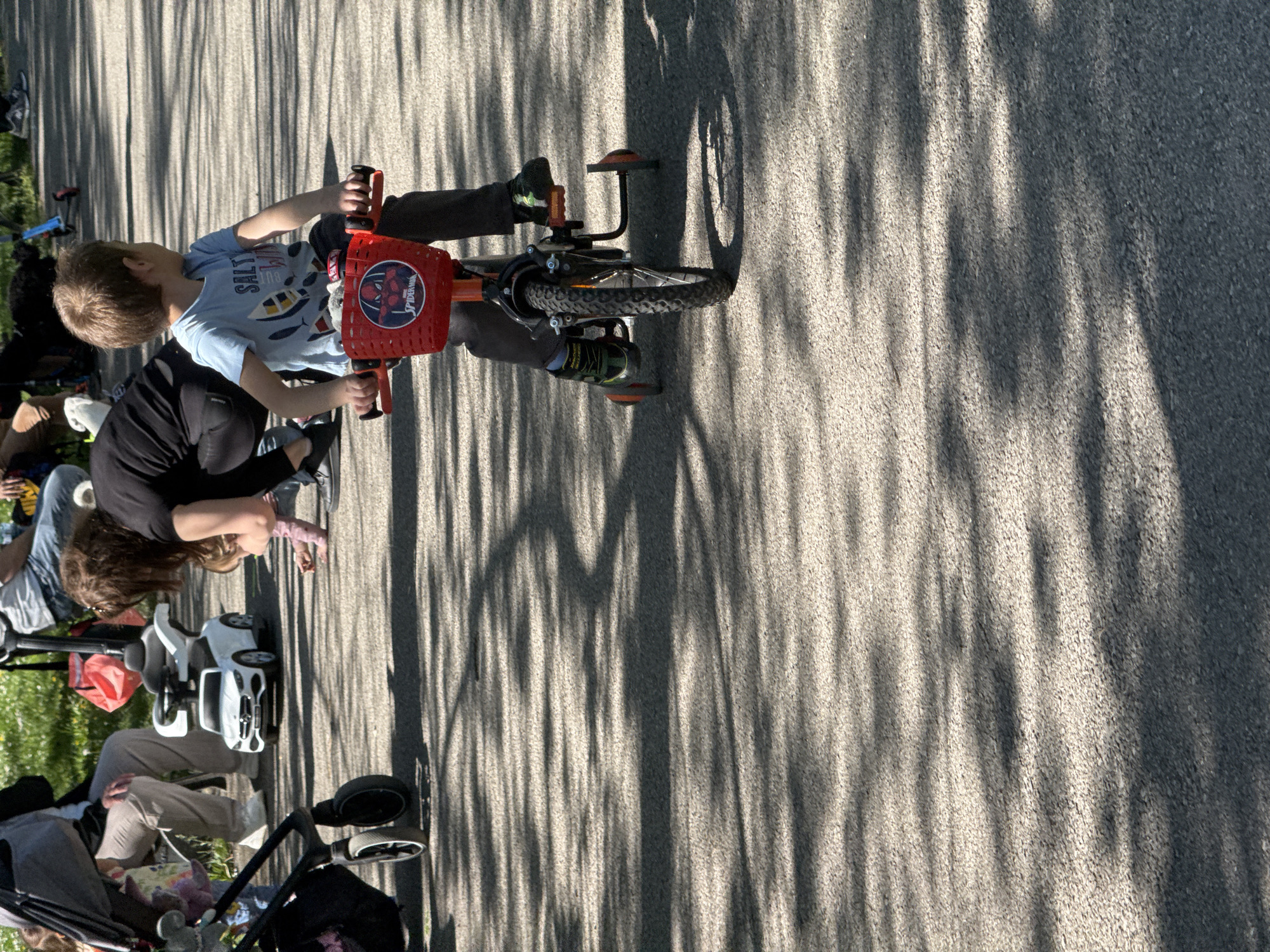 A photograph shows a child on a Spider-Man bicycle on a paved road. The child wears a light blue t-shirt with a picture of fish. Another person stands next to them. A stroller and a white toy car are visible on the grass near the road, suggesting a park setting. Tree shadows are projected on the pavement. The precise location in Sofia, Bulgaria, cannot be determined from this image alone.