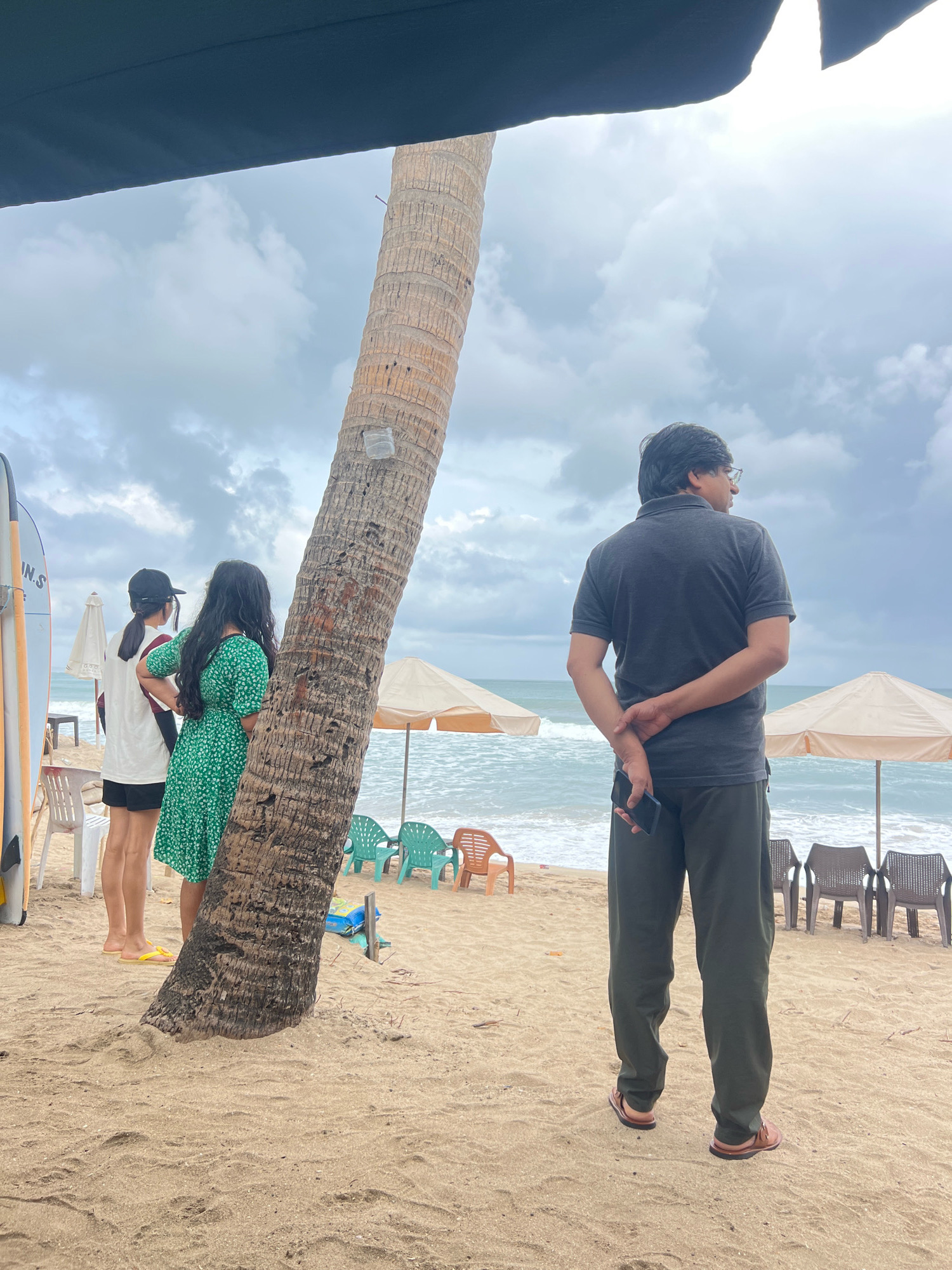 On a cloudy day at Kuta Beach, Indonesia, four individuals stand on the sandy shore. A man in a dark grey polo shirt and dark green pants stands with his back to the camera, holding a black smartphone in his right hand, his arms clasped behind him. In front of him and to his left, a woman in a short-sleeved green floral dress stands facing away, her hands on her hips. Next to her, a person wearing a white t-shirt, black shorts, and a black baseball cap also faces away. To the far left, a surfboard stands upright. Several plastic chairs in green and orange, along with beach umbrellas, are scattered on the sand facing the ocean, where waves are visible. A thick palm tree trunk stands prominently in the foreground.