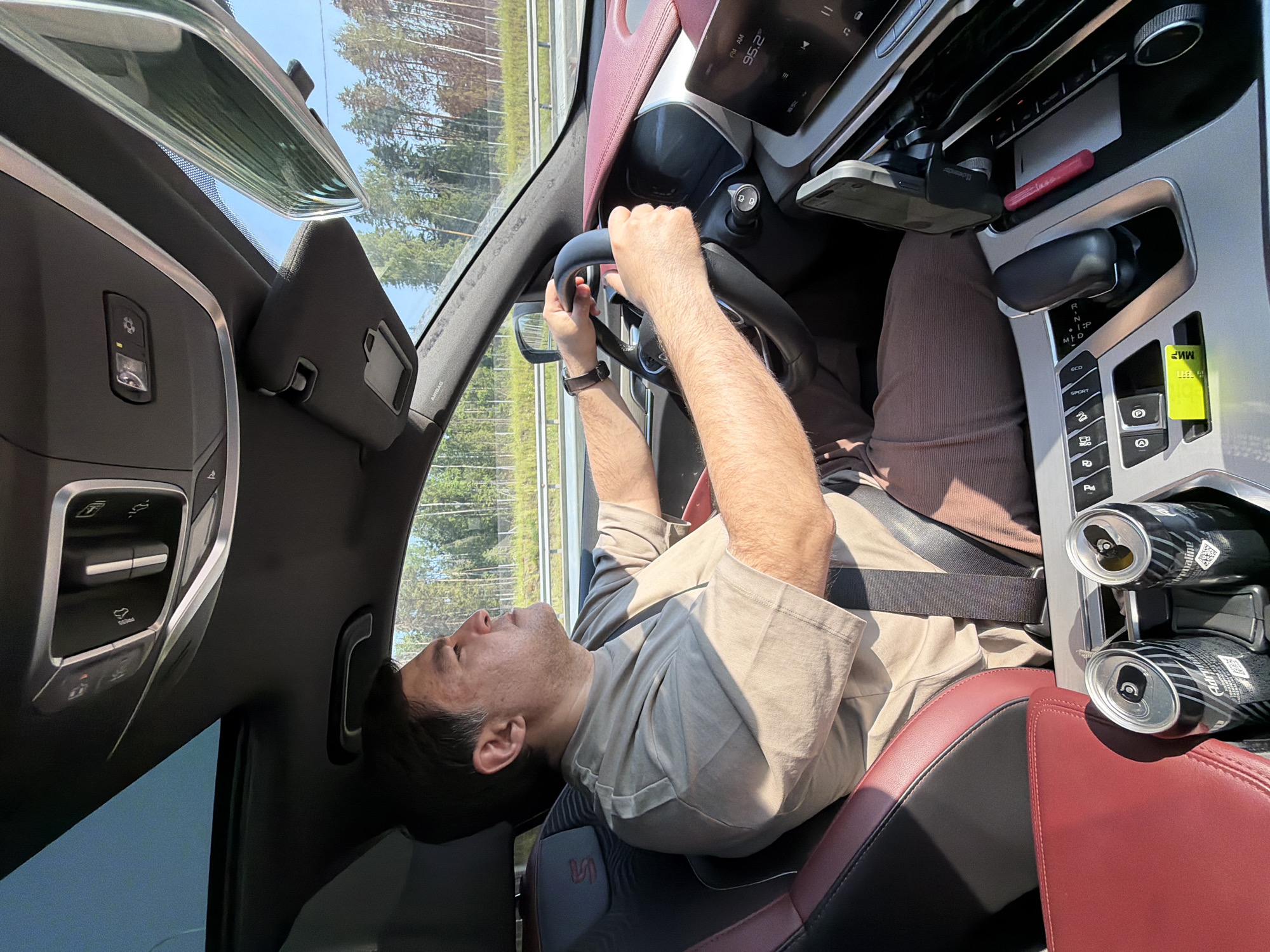 An adult male is driving a car. He is wearing a tan t-shirt and brown pants. He has his hands on the steering wheel. The car has a black interior with red accents. There are two cans of energy drink in the cup holder. The car is driving on a highway with trees visible on the side. The sun is shining.
