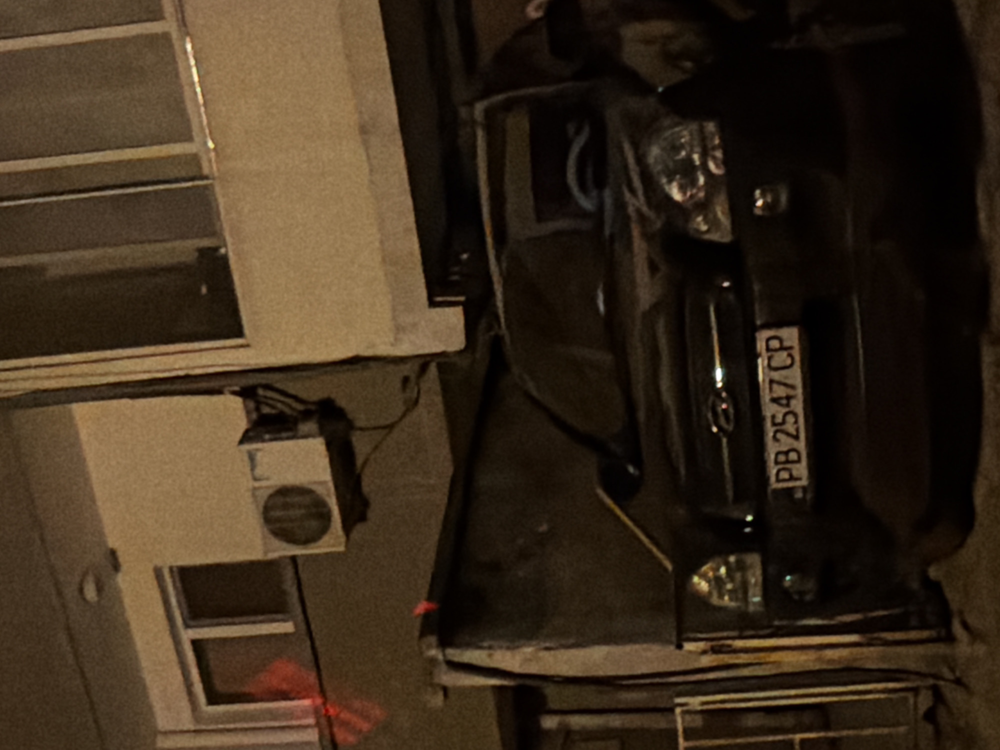 The image shows a dark-colored car with a license plate reading "PB 2547 CP". The car is parked in front of a building with an outdoor air conditioning unit mounted on the wall. The scene appears to be at night, with artificial lighting casting shadows and illuminating parts of the car and building. The area is urban, likely in Plovdiv, Bulgaria, given the license plate format. A partially visible metal fence is to the left of the car. There are no people visible in the image. The car's front end is visible, including its headlights and grill.