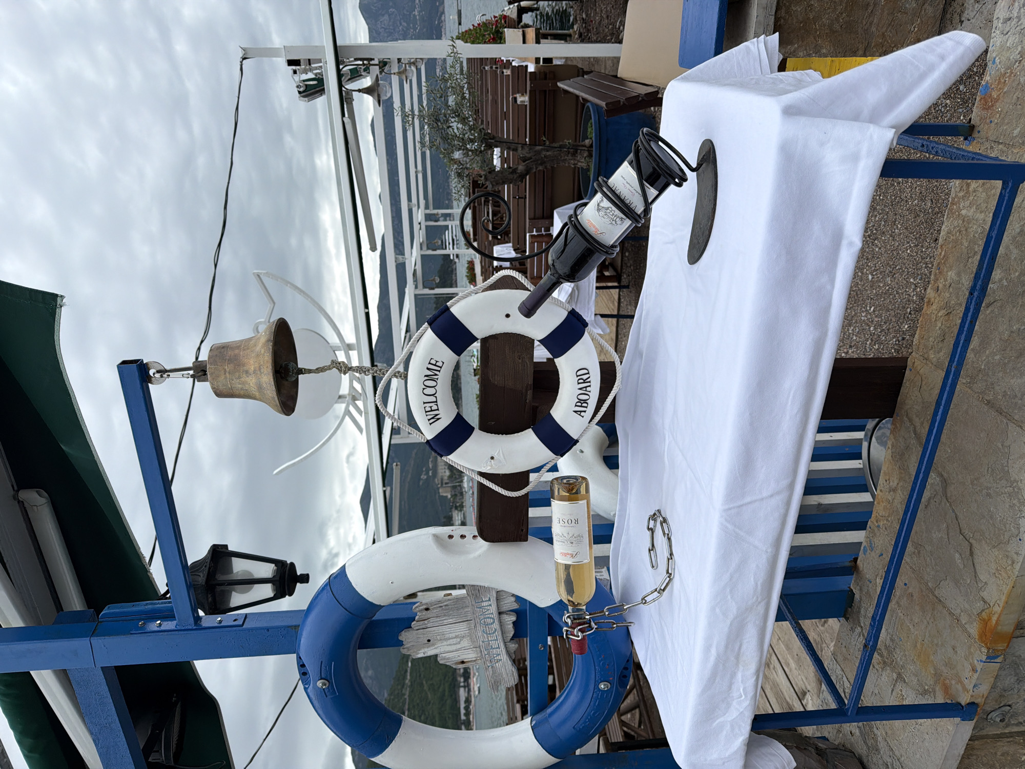 The image shows a decorative nautical setup, likely on a boat or at a waterfront location. There are two life rings, one white with blue stripes and another white with the words "WELCOME ABOARD" in blue. A brass bell hangs from a blue painted metal frame, along with an ornamental lantern. A white wine bottle is secured with a chain, and a bottle of red wine is suspended upside down in a decorative holder. The background shows buildings and landscapes. Based on the context of this task, it is plausible to believe this might be in Budva, Montenegro.