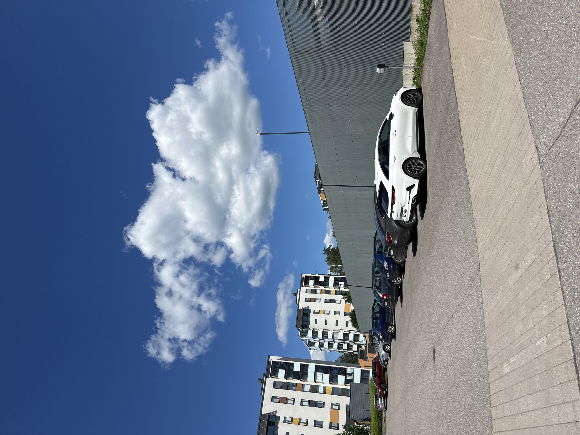 A wide, eye-level, outdoor shot captures a parking lot in front of modern apartment buildings and a gray corrugated metal fence. The sky is a vibrant blue, with a large, puffy white cloud dominating the upper center of the frame. In the foreground, the asphalt parking lot is lined with several cars, including a white Kia station wagon parked on the right. Beyond the parking lot, two multi-story apartment buildings with white facades and colorful balconies stand to the left. A tall, gray corrugated metal fence runs along the middle of the frame, separating the parking lot from an unseen area. A lamppost with a light fixture is visible in the distance, extending above the fence. The scene is bathed in bright sunlight, casting distinct shadows.
