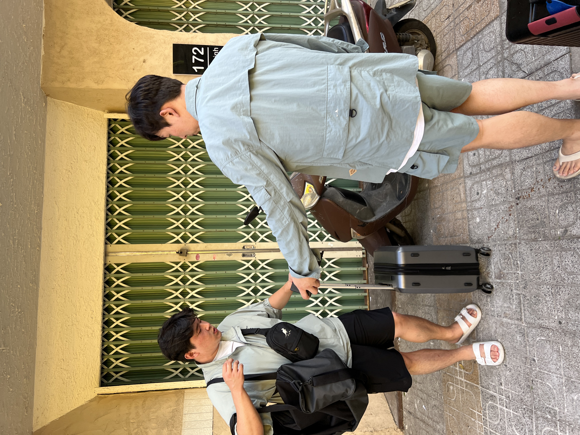 Two adult males, appearing to be of East Asian descent, are standing on a paved sidewalk in front of a building with a pale yellow facade and green metal security gates. The man on the left is facing towards the viewer and is carrying two black bags, one over his shoulder and one in his left hand. He is wearing a light green short-sleeved jacket over a white t-shirt and black shorts. He has white slide sandals on his feet. The man on the right is facing away from the viewer and is wearing a light blue long-sleeved jacket and matching shorts. He is holding a dark object in his right hand and is reaching towards a grey rolling suitcase positioned on the sidewalk between the two men. Behind them, a brown scooter is parked. A building number "172" is visible on the wall to the right of the security gates. The overall scene suggests a departure or arrival, possibly related to travel. The location is identified as Nha Trang, Vietnam.