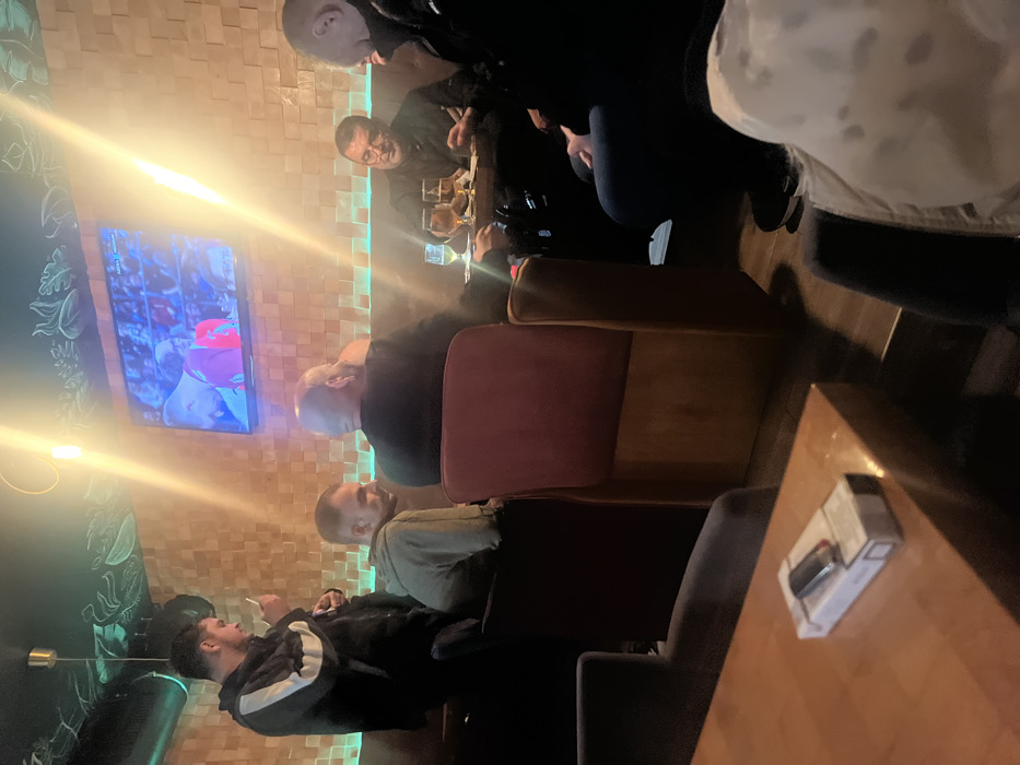 A group of men gather around a table in a bar. Some are watching a sporting event on a nearby TV. A man in a black jacket with white stripes smokes a cigarette. A lighter is on the table. The bar has a textured wall with green and blue neon lighting.
