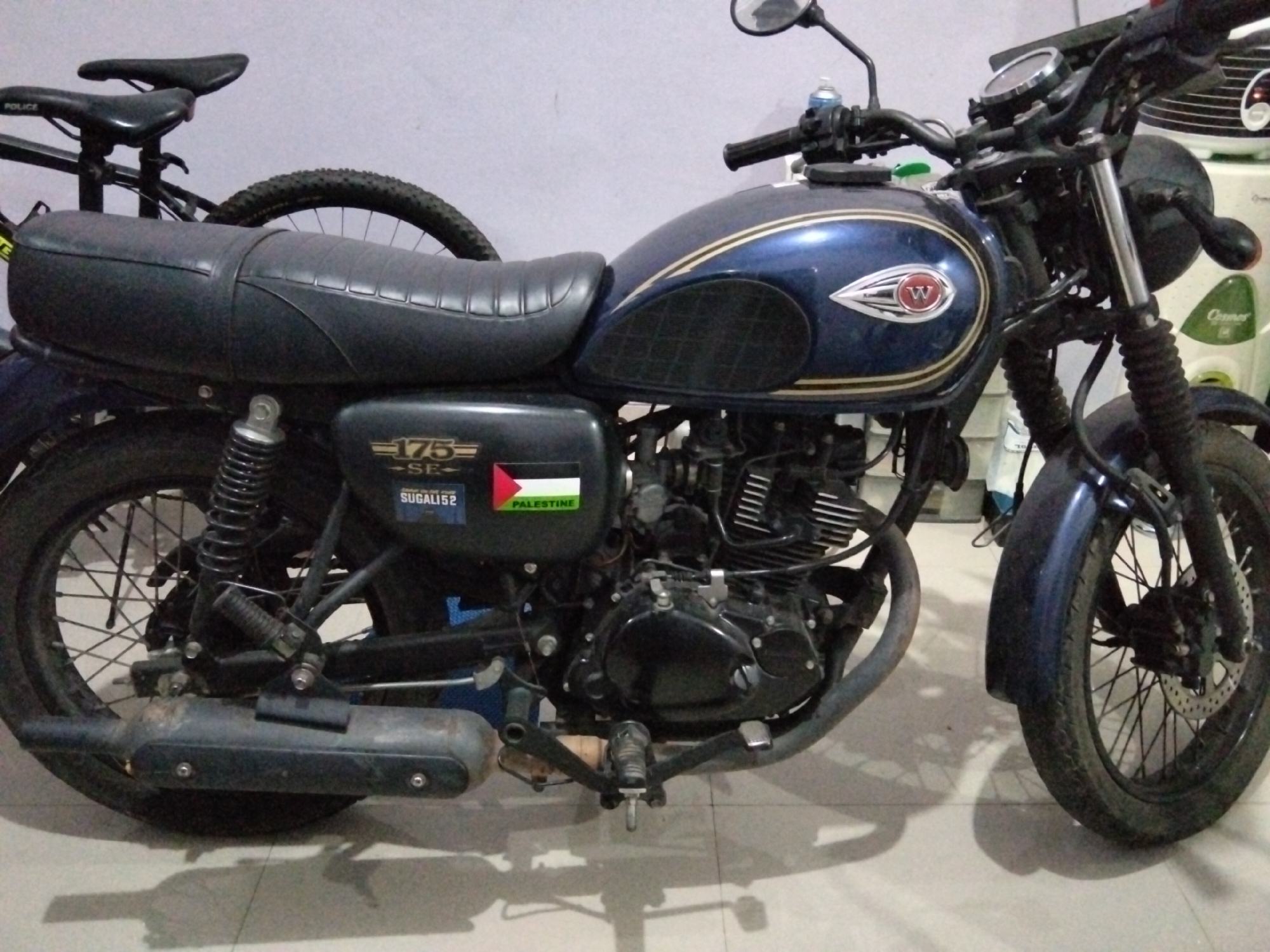 The image shows a dark blue motorcycle parked indoors, possibly in a garage or a room. The motorcycle is the primary subject and is viewed from the side. It has a black seat, black handlebars, and a black exhaust pipe. There are visible stickers on the side of the motorcycle, one appearing to be the flag of Palestine. The motorcycle's engine is visible. In the background, a white appliance, likely a washing machine or refrigerator, is partially visible. There's also a glimpse of another bicycle behind the motorcycle. The lighting suggests an indoor environment, with shadows cast on the tiled floor. There are no people visible in the image. The image is taken in Rawa Atug, Indonesia.