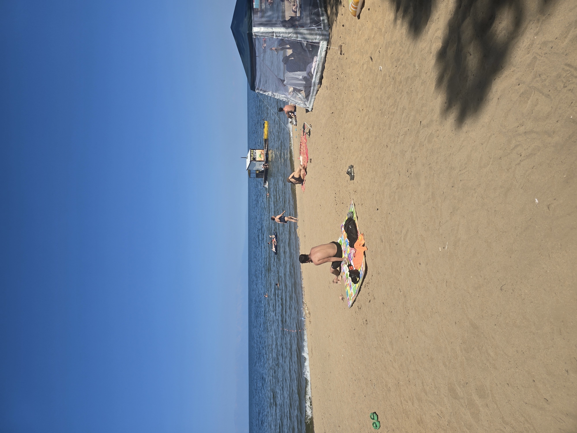 A sunny day at a sandy beach on the shore of a large body of water, possibly a lake or sea, with a clear blue sky. In the foreground, a man sits on a colorful beach towel, facing away from the camera, looking out at the water. To his left, a pair of green flip-flops are placed on the sand. In the middle ground, several people are visible in the water, some swimming and others on inflatable toys. Further back, a floating structure with a small building on it is anchored in the water. To the right of the image, a large, transparent tent provides shade for several people. The beach is mostly empty, with a few other individuals scattered around. The water is calm with small waves lapping at the shore. The overall atmosphere is one of relaxation and leisure on a warm day. The location is Maksimiha, Russia.