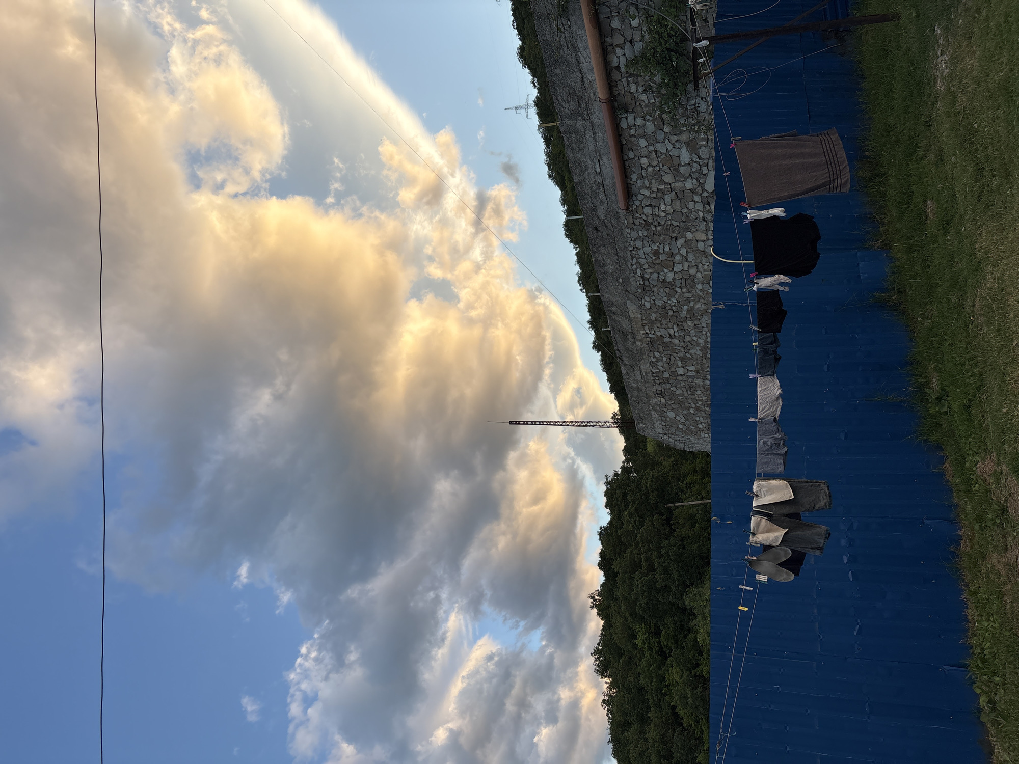 This is a low-angle outdoor photograph taken in the city of Chaika, Ukraine. The scene is dominated by a bright blue metal fence in the foreground. Clothes, including underwear and towels, are hanging on a clothesline strung across the fence, secured by clothespins. The grass in front of the fence is green and somewhat overgrown.
Behind the fence, a stone wall rises, topped by a dense line of green trees that cover a hillside. A dark, lattice-like tower is visible on top of the hill in the distance, along with what appears to be electrical pylons.
The sky above is dramatic, with a mix of large, fluffy clouds. Some of the clouds are illuminated by the setting sun, casting a warm, golden glow, while others are darker gray. A thin wire, possibly for electricity or communication, is stretched across the upper portion of the frame. The overall impression is one of a rural or semi-rural setting during late afternoon or early evening.