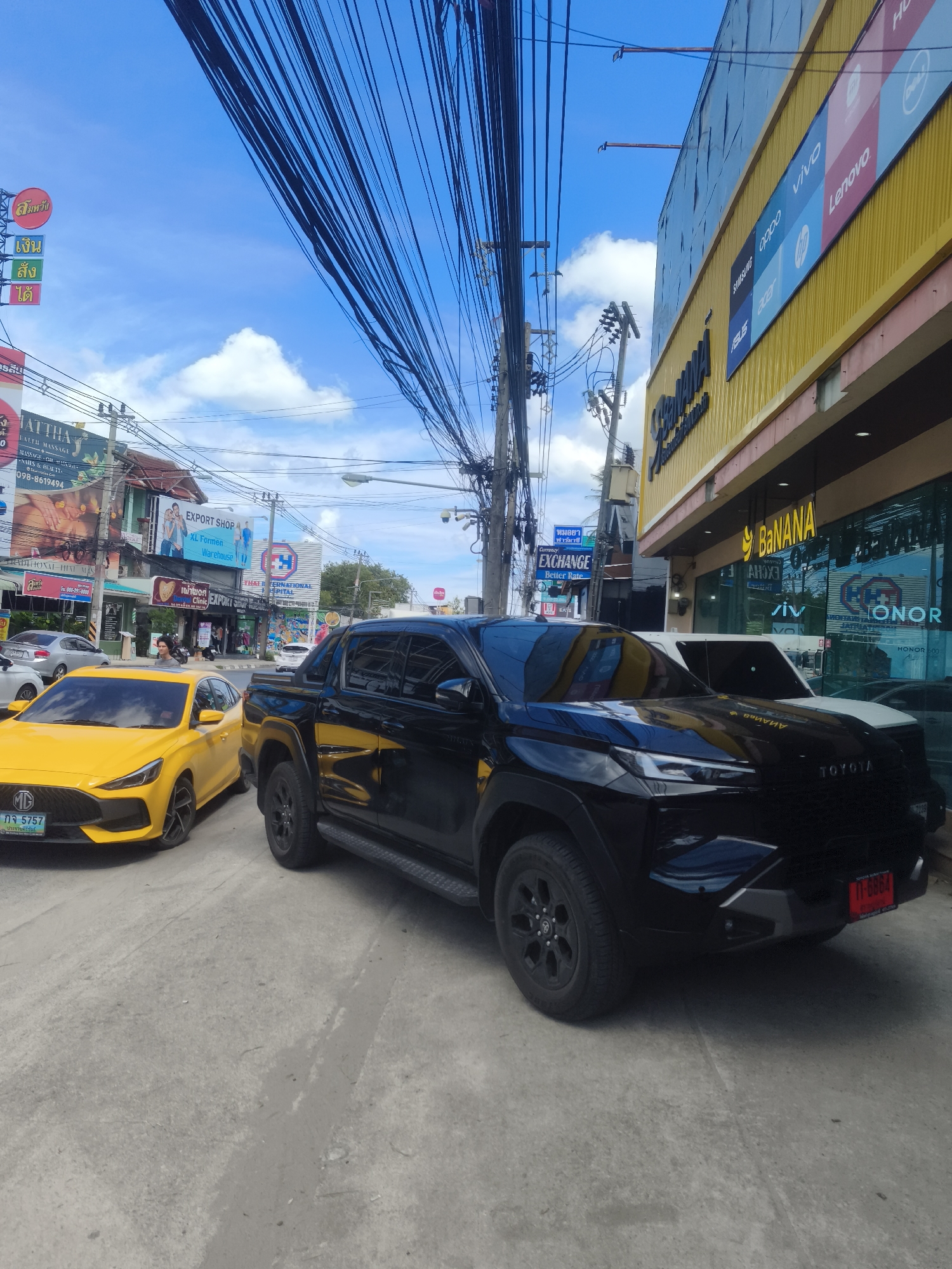 The image captures a street scene in the Ko Samui District of Thailand. In the foreground, a black Toyota pickup truck is parked on a concrete road. To its left, a bright yellow MG sedan is also parked. The road is lined with buildings displaying various signs and advertisements. One building, painted yellow, features large lettering that reads "BANANA" and displays logos for electronic brands such as Vivo, Lenovo, and Honor. Another building has signs for "EXPORT SHOP" and "THAI INTERNATIONAL HOSPITAL". The sky above is blue with scattered white clouds, and a dense network of electrical wires is visible overhead. The environment appears to be an urban commercial area with shops and businesses.