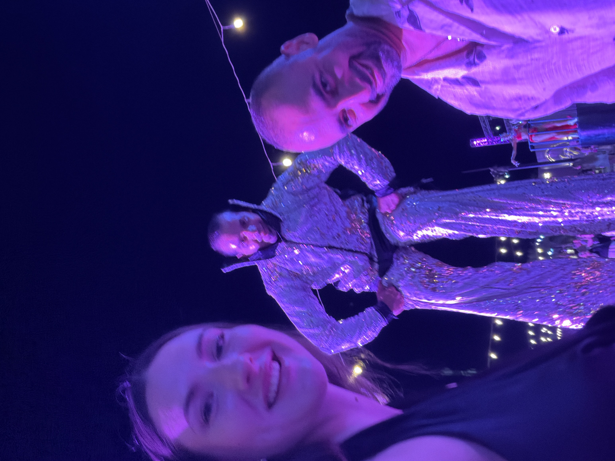 A group of people are gathered outdoors at night, possibly at an event or festival in Sharm El Sheikh, Egypt. The scene is illuminated by purple lights, with strings of yellow bulbs visible in the background. In the foreground, a woman with long brown hair smiles towards the camera. To her right, a man with a bald head and a beard grins, wearing a light-colored patterned shirt. Further back, a performer on stilts is dressed in a sparkly, sequined outfit that reflects the purple lighting. The performer's arms are bent, and they are looking down. Another individual in a red costume is visible on a platform in the distance.