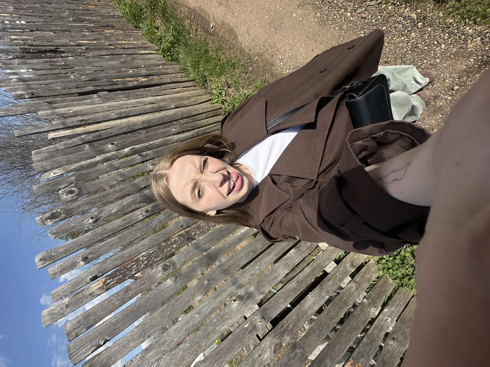 A selfie taken in an outdoor setting, possibly in or near the city of Chaikovsky, Russia, showing a young woman with fair skin, blonde hair, and braces on her teeth. She is wearing a brown trench coat over a white top and is looking at the camera with a somewhat playful expression, sticking her tongue out slightly. A black handbag is visible near her. The background is dominated by a weathered wooden fence with patches of grass and dirt. A small portion of the sky is visible in the upper left corner of the frame. The selfie angle is slightly tilted. Part of the person's arm is visible on the right side of the picture.