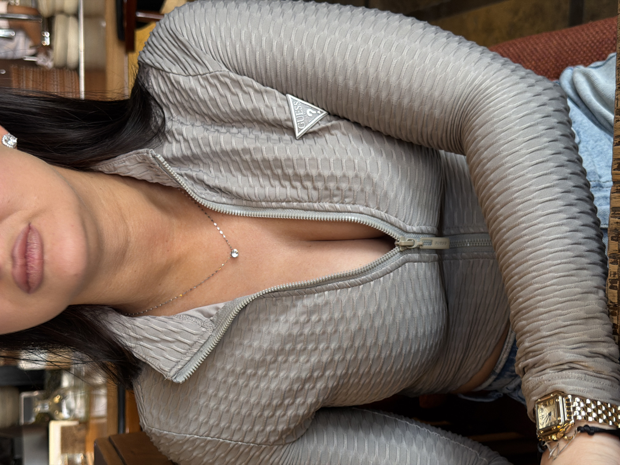 A woman is wearing a gray textured jacket with a "Guess" logo and a silver necklace with a small gemstone. She is also wearing a gold wristwatch. The background appears to be a restaurant or cafe setting. There is no indication that the location is in Moscow, Russia.