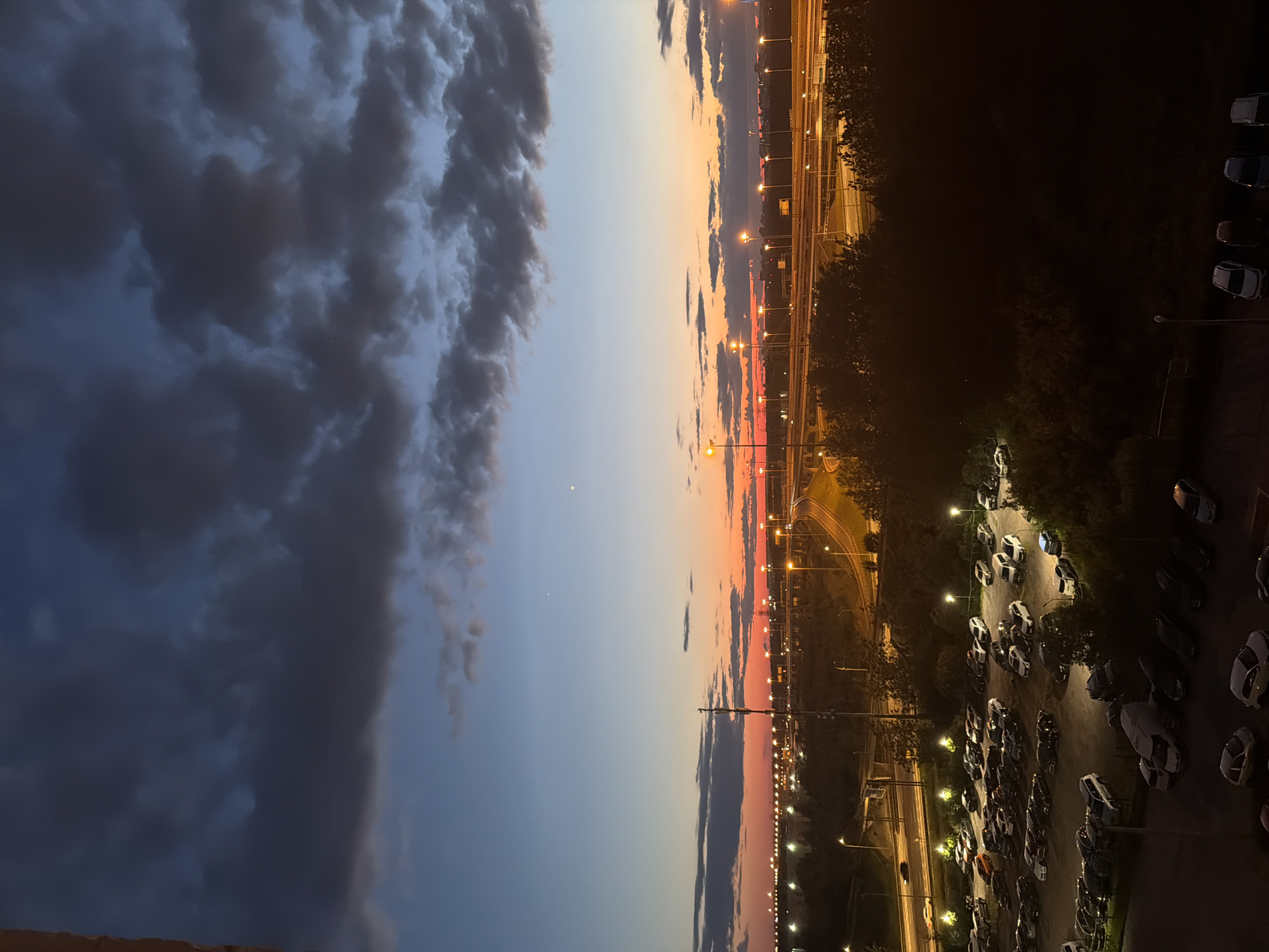 The image depicts an outdoor scene at dusk, likely in an urban or suburban environment. The sky is a gradient of blues and oranges, with dark clouds accumulating towards the top. In the midground, a highway system is visible, illuminated by streetlights and the fading light of the sunset. Numerous cars are parked in a large lot in the foreground and midground, some with their headlights on. Trees and other vegetation are scattered throughout the scene, casting shadows in the low light. The overall impression is one of a transition from day to night, with artificial lights beginning to dominate the landscape.