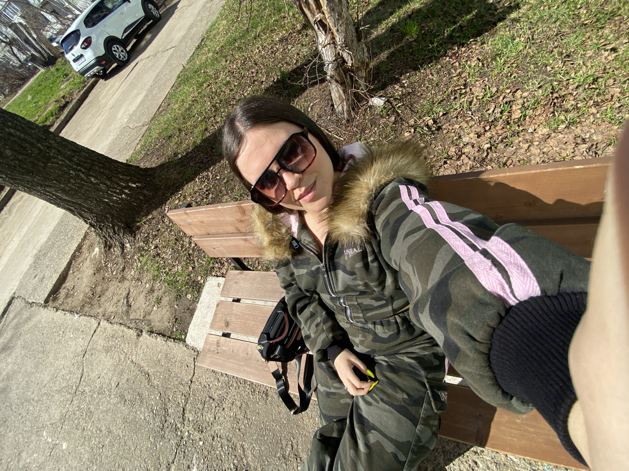 The image, likely a selfie, shows a woman wearing sunglasses, a camouflage tracksuit with pink stripes, and a fur-lined hood. She is sitting on a wooden bench, with a black backpack next to her. A tree trunk is visible to her left, with grass and fallen leaves in the background. A concrete sidewalk is also visible.
In the upper left corner of the image, a white car is parked on a street. Based on the style of buildings and the vegetation, the location is identified as the city of Chaikovsky, Russia. The woman appears to be relaxing on the bench, possibly taking a break. Her right hand is in the foreground.