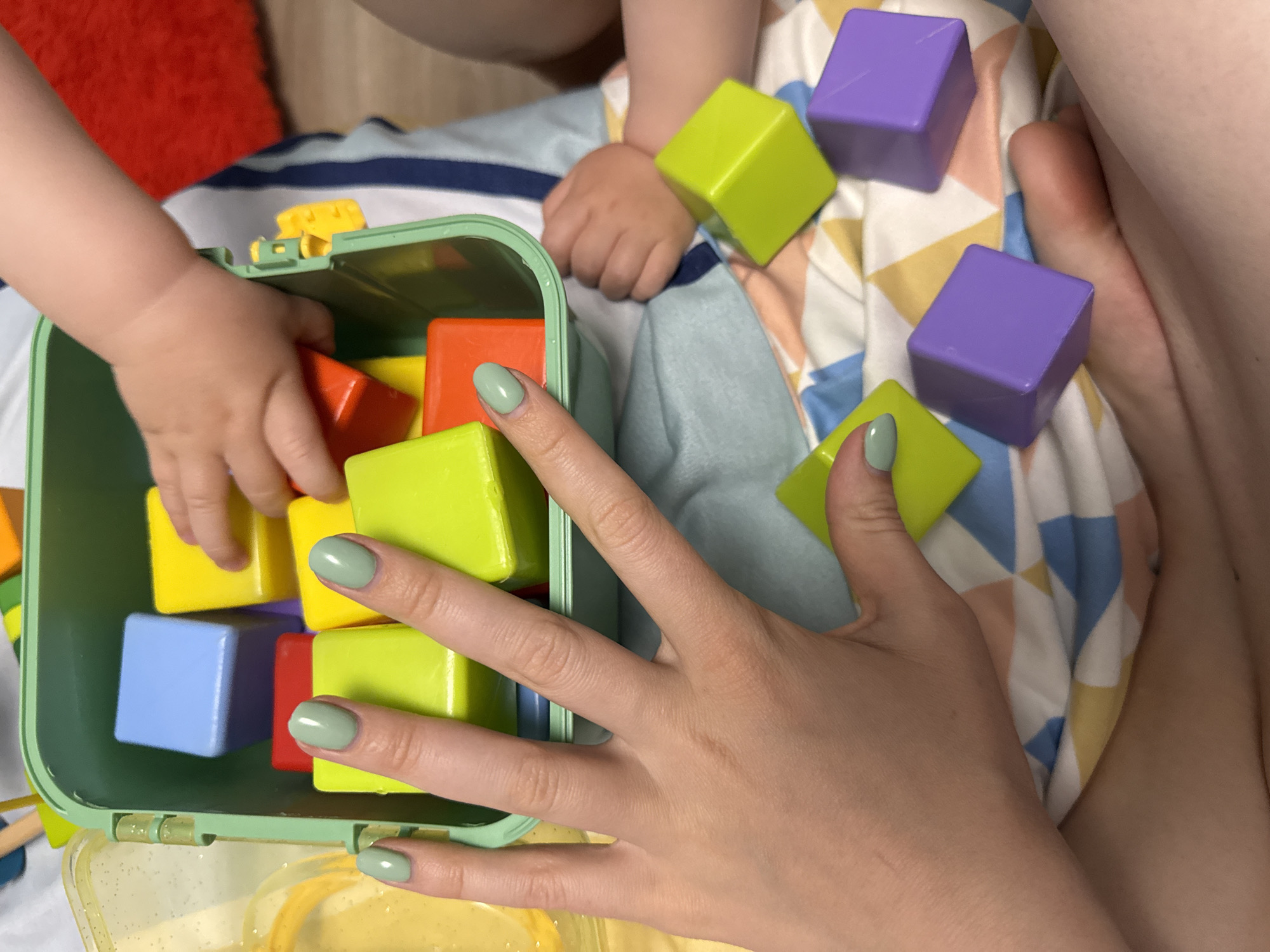 The image shows an aerial view of a child and an adult playing with colorful blocks on a patterned cloth. The child's hand is inside a green plastic container filled with blocks of various colors (yellow, orange, blue, and green). Several blocks are scattered on the cloth surface: green and purple. The adult's hand, with light green nail polish, rests among the blocks. The cloth has a geometric pattern with colors like blue, white, yellow, and pink. An out-of-focus red cushion is visible in the upper left corner. The setting appears to be indoors. There is no evidence to connect this scene to the city of Moscow, Russia.