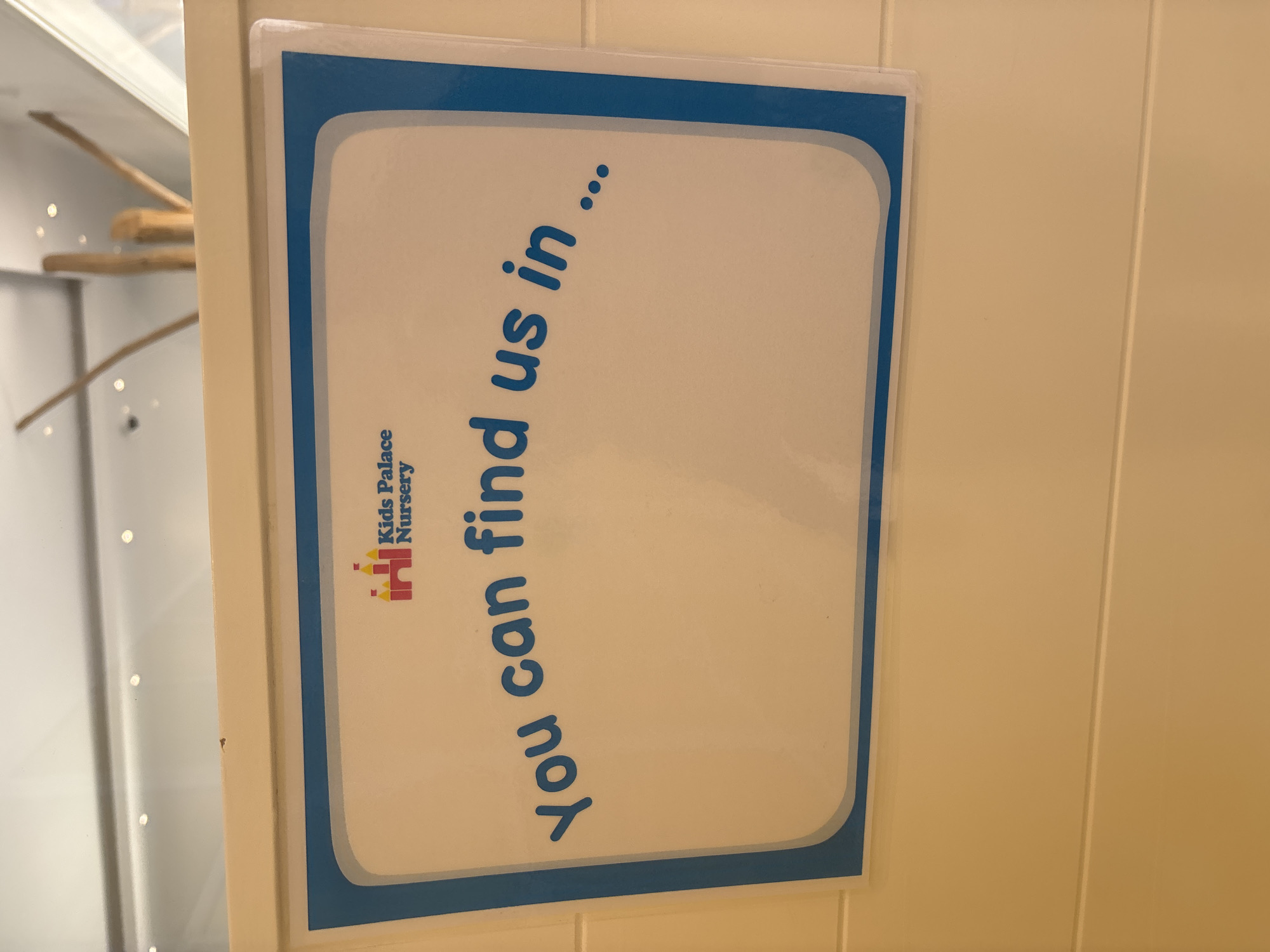 The image shows a close-up of a sign that reads "You can find us in..." with the logo for "Kids Palace Nursery" above the text. The sign is framed with a blue border and is affixed to a light-colored wall with horizontal paneling. The background is blurred but appears to be an indoor setting with some wooden structures and ceiling lights. There are no people or animals visible in the image. The sign is the primary subject, and its placement suggests it is intended to inform visitors about the location of the nursery. The text and logo are in a playful, child-friendly style. There is no information in the image that allows for the determination of its location in Dubai, United Arab Emirates.