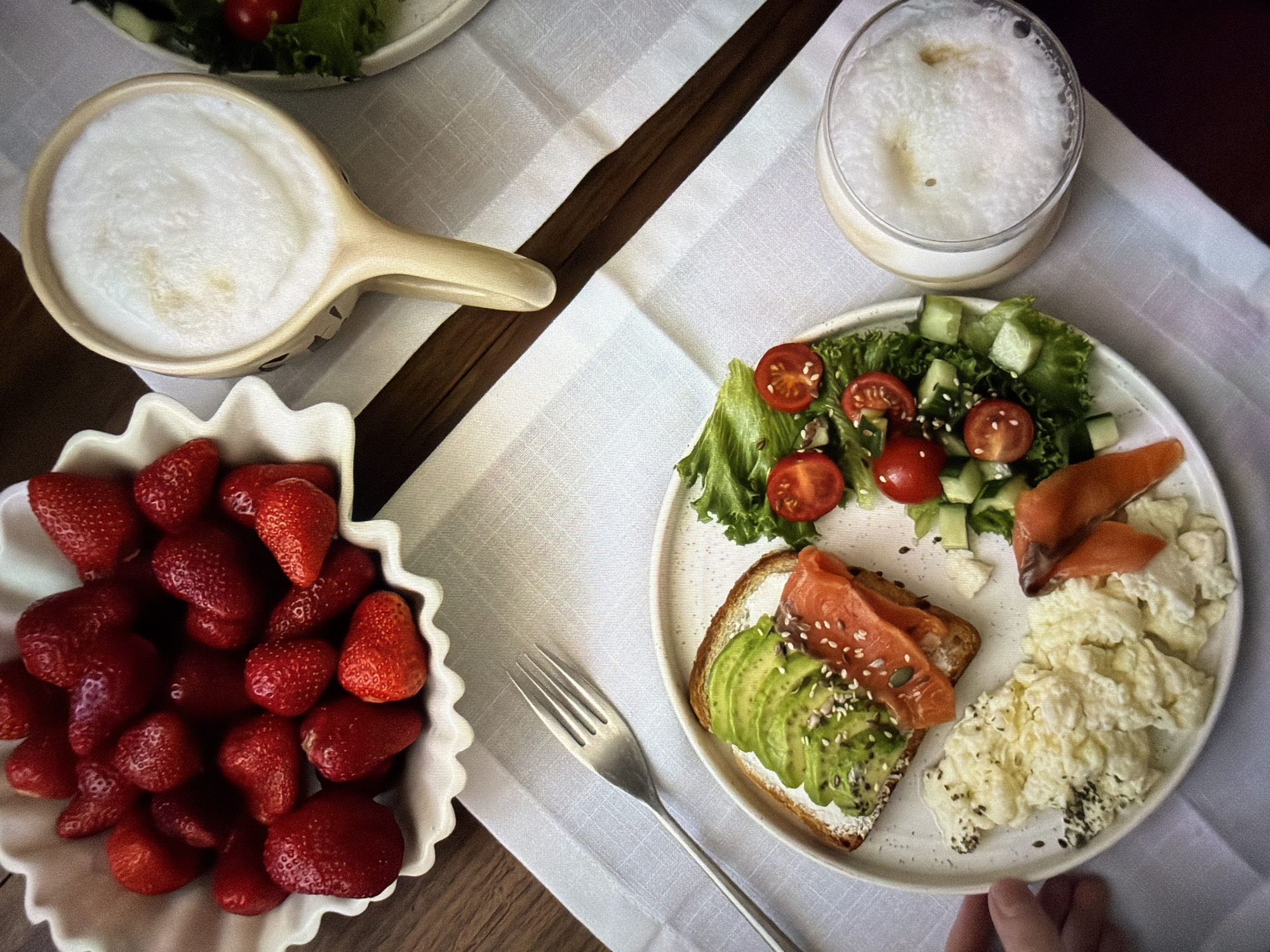 An overhead shot captures a beautifully arranged breakfast or brunch spread on a light-colored, textured tablecloth, set upon a dark wooden table. A central white ceramic plate holds a diverse meal: a slice of toasted bread topped with sliced avocado, a white spread (likely cream cheese), and various seeds (sesame, flax, poppy, or similar). Beside the toast are several slices of smoked salmon and a generous portion of fluffy scrambled eggs, also sprinkled with dark seeds. A small side salad of green lettuce, halved cherry tomatoes, and diced cucumber completes the plate.
To the left, a white ceramic bowl with a decorative wavy rim is brimming with fresh, ripe red strawberries. Two frothy beverages are visible: one in a light-colored ceramic mug or pitcher with a handle, and another in a clear glass, both topped with a thick layer of white foam, suggesting coffee or milk-based drinks like lattes or cappuccinos. A silver metal fork rests on the tablecloth, pointing towards the main plate. A hand, likely adult, is subtly visible in the bottom right corner, suggesting interaction with the table setting. The overall presentation is neat and appealing, indicative of a leisurely meal enjoyed during the daytime. No specific location cues within the image point to Ros Na Rí, Ireland, other than the provided context. There is no visible text in the image.