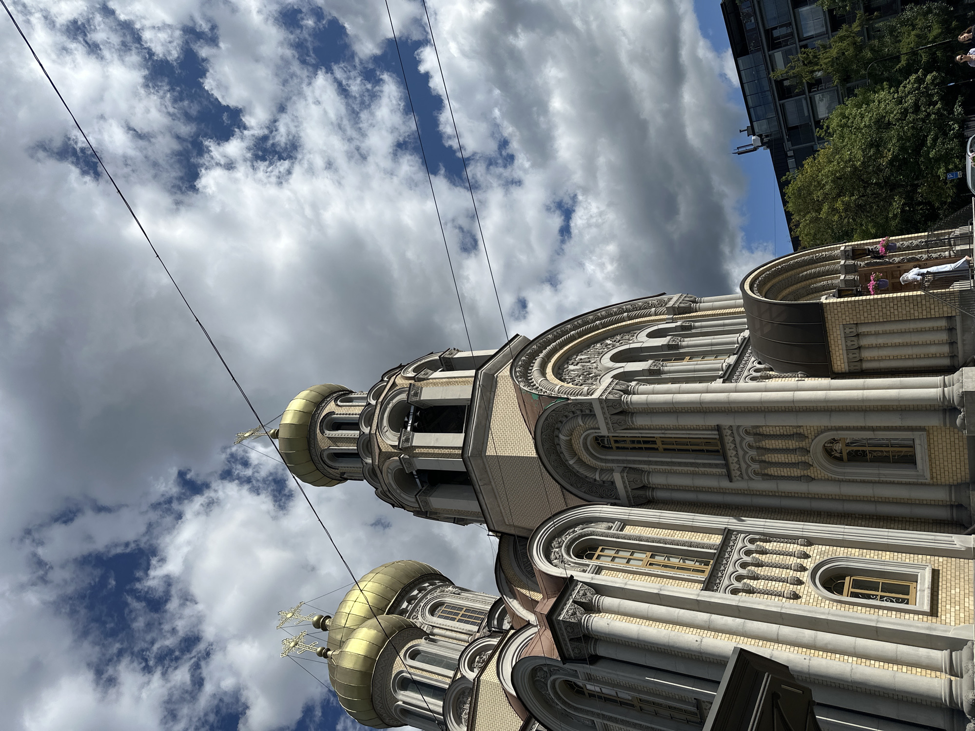 The image shows the Resurrection Church in Vilnius, Lithuania, an Orthodox church with distinctive golden domes topped with crosses. The church's facade is adorned with yellow brickwork and grey columns and intricate architectural details. Two golden domes are prominent in the foreground, with a third partially visible. The sky above is dramatic, filled with a mix of dark and light clouds, with patches of blue sky visible. Two thin black wires stretch across the upper portion of the image. To the right of the church, a modern building with balconies and trees can be seen. A person dressed in white is standing on the steps of the church entrance. The scene appears to be captured from a low angle, looking up towards the church and the sky.