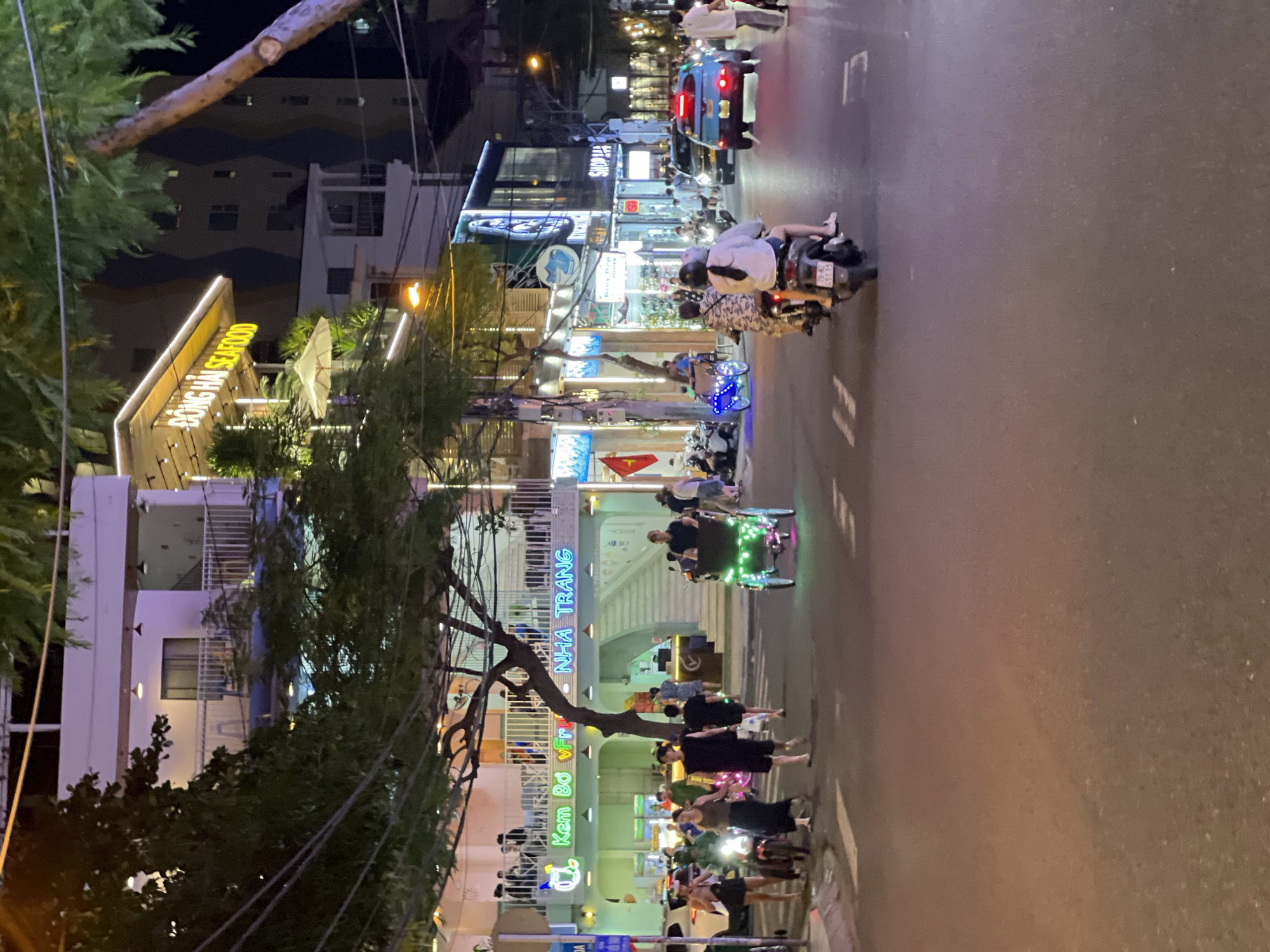 This is a nighttime street scene in Nha Trang, Vietnam. A two-story building with a lit-up sign reading "DONG HAI SEAFOOD" is visible on the right side of the image. To the left, another building has a bright green facade with signs reading "Kem Bo v Fruit" and "NHA TRANG." People are walking on the sidewalk and crossing the street. Several motorbikes are present, with some individuals riding them. One illuminated rickshaw with neon lights is also on the street. Cars and other vehicles are driving on the road, which is illuminated by streetlights and the lights from the buildings. The overall atmosphere is bustling and active, typical of a commercial street at night in Vietnam.