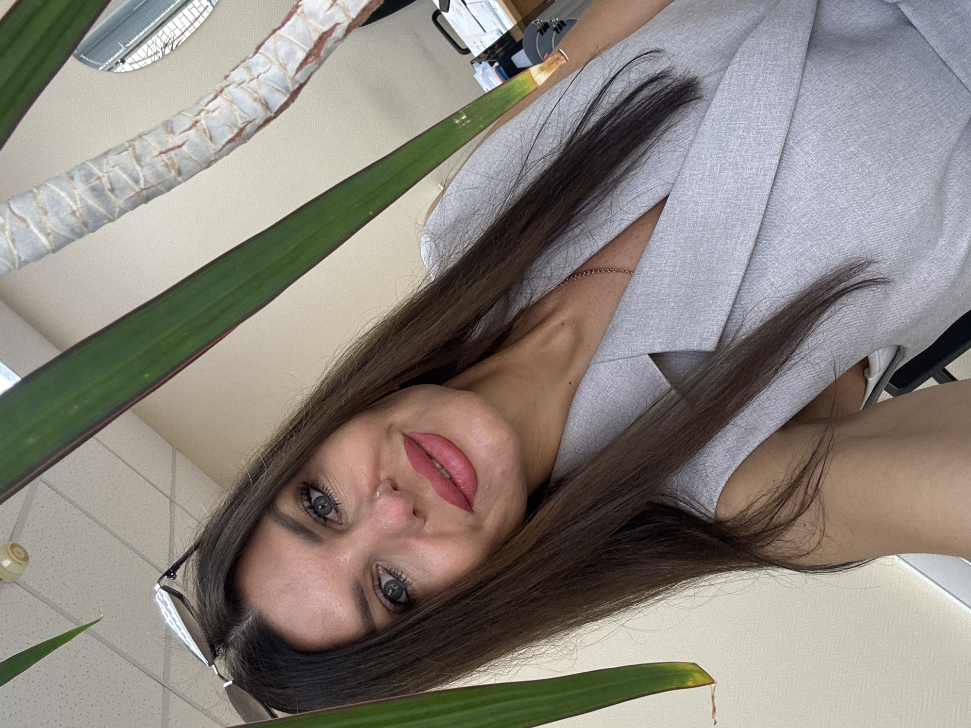 A woman with long, dark brown hair and blue eyes is taking a selfie. She is wearing a light gray sleeveless blazer. Her makeup includes pink lipstick and eye makeup. Sunglasses are perched on top of her head. She has a small nose ring. The background is an indoor setting, with a light-colored wall and ceiling. A ceiling light fixture is visible. Part of a large plant with green leaves and a textured stalk is in the foreground, framing the woman's face. In the background, to the right, a desk with papers and what appears to be a computer monitor is visible, suggesting an office environment. The image does not contain any elements that would allow for identification of the location in Chaikovsky, Russia.