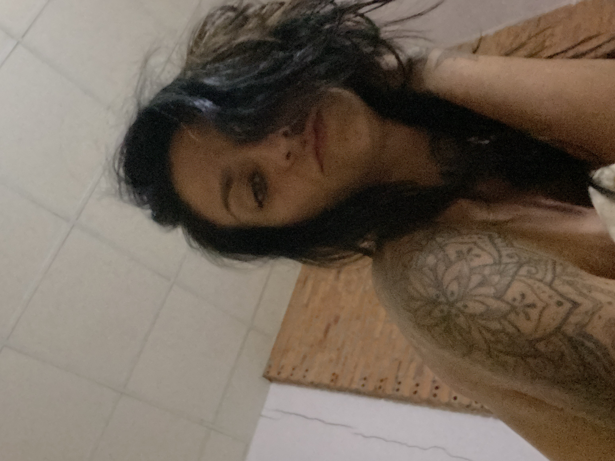 A low-light, upward-angled portrait features a female individual indoors. She has dark hair, with a portion falling across the right side of her face, and her gaze is directed straight into the camera. A prominent, intricate mandala-patterned tattoo is visible on her left shoulder and upper arm. The individual appears to be bare-shouldered or wearing minimal upper body attire. The background consists of a light-colored ceiling with grid lines. To the left, a white wall shows distinct vertical cracks. Further left, a wooden structure made of light-colored vertical planks with small, dark, regularly spaced circular marks (likely fasteners) is visible. The lighting is dim, contributing to a slightly grainy image quality. This scene is situated in Mũi Né, Vietnam.