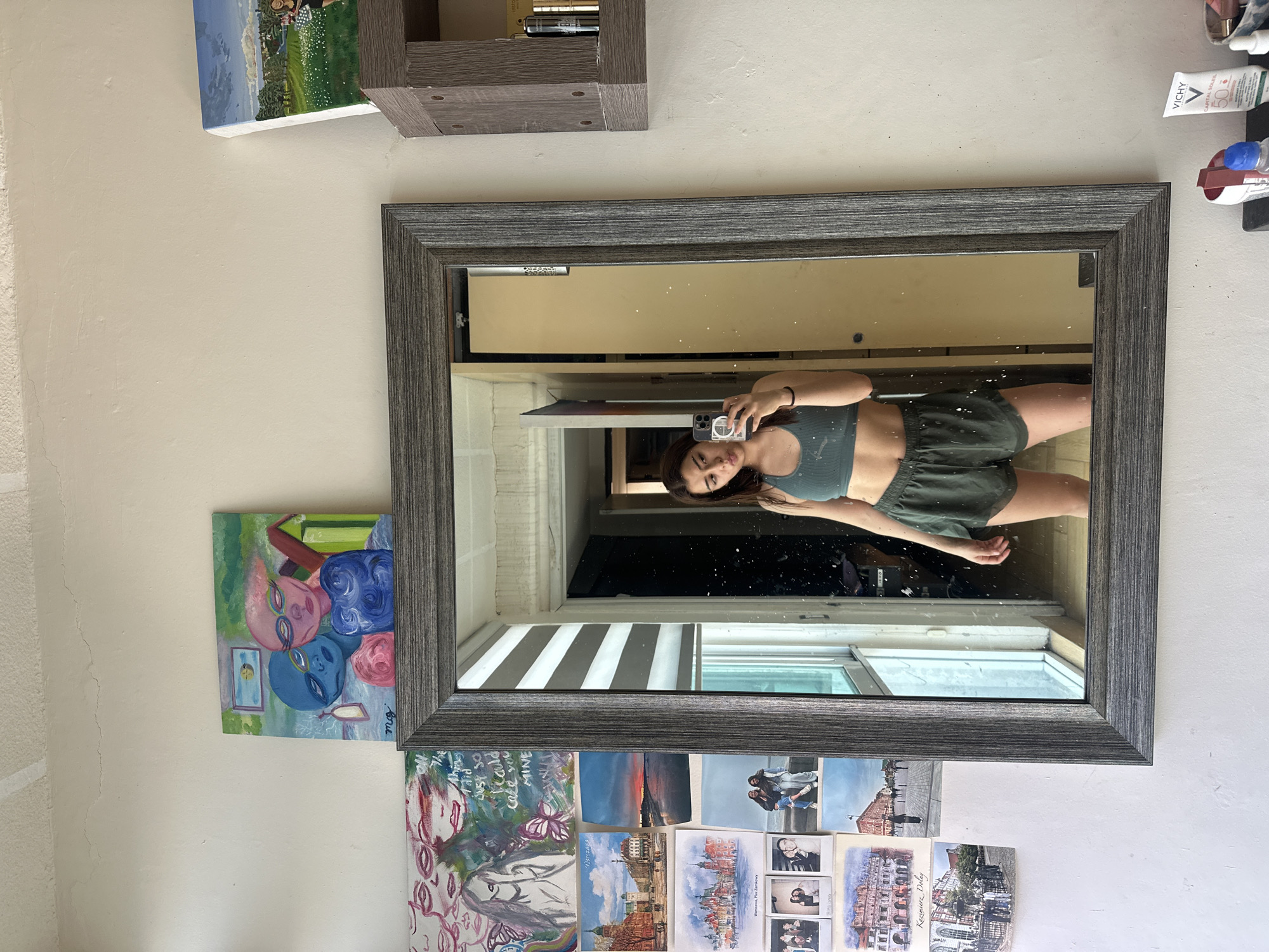 A mirror, framed in grey wood, reflects a person holding a cell phone in their hand, taking a selfie. The person is wearing a dark green athletic top and shorts. The mirror is mounted on a light tan wall that features a variety of other objects: a small grey shelf, a painting with a green field and people, an abstract painting featuring various pink, red, and blue alien-like faces, and a collection of postcards. The postcards include images of skylines, landscapes, and people. The reflection in the mirror shows a partially-open window with white Venetian blinds, and a hallway with a door on the left.