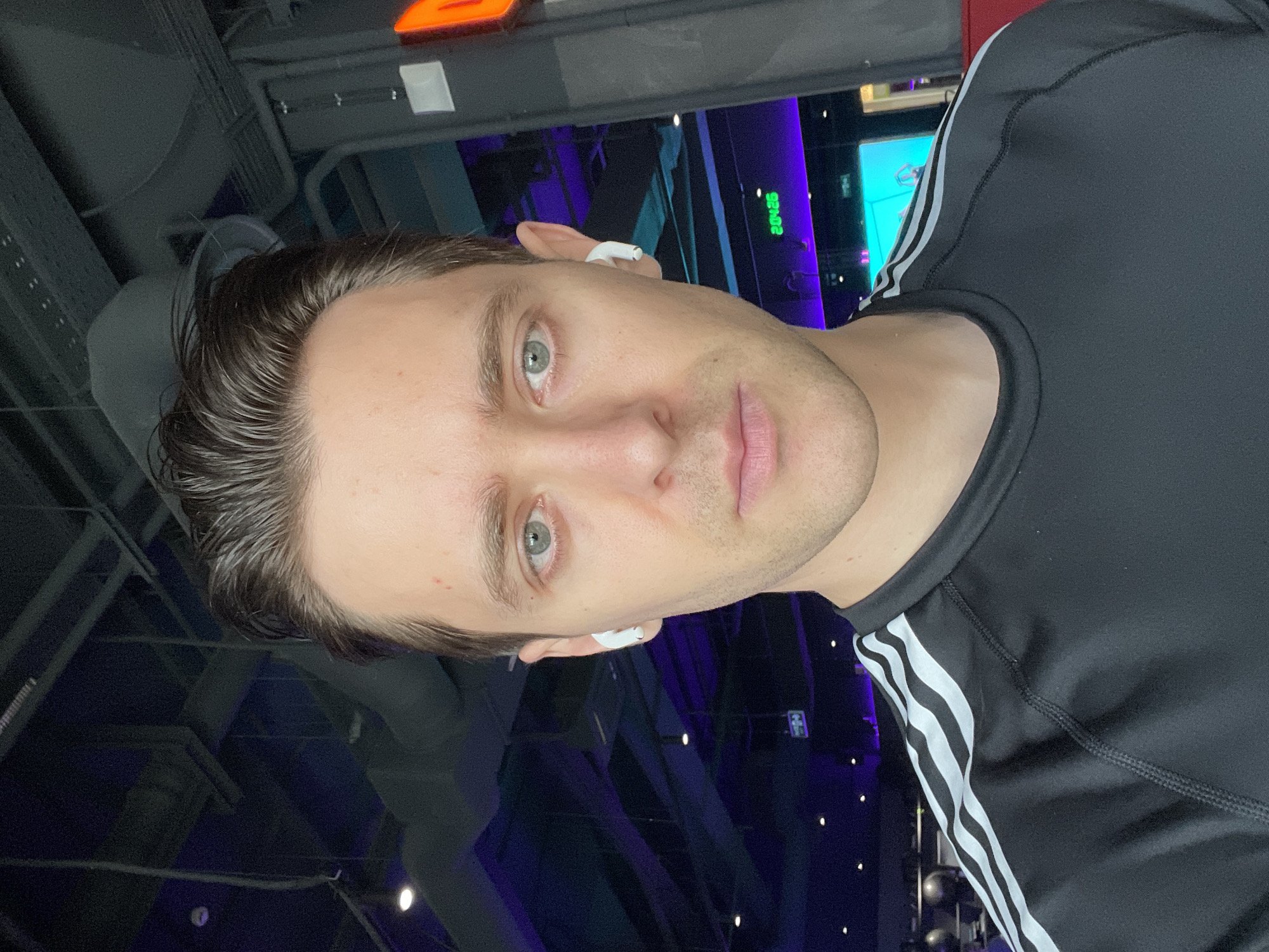 A close-up indoor shot depicts a light-skinned male with short, dark hair and light-colored eyes. He is wearing a black athletic shirt with white stripes on the shoulder. He has wireless earbuds in both ears. The background features a partially visible area with blue and purple lighting, appearing to be some sort of arena or stage. The location is potentially Saint Petersburg, Russia, but without further context, this cannot be definitively confirmed.