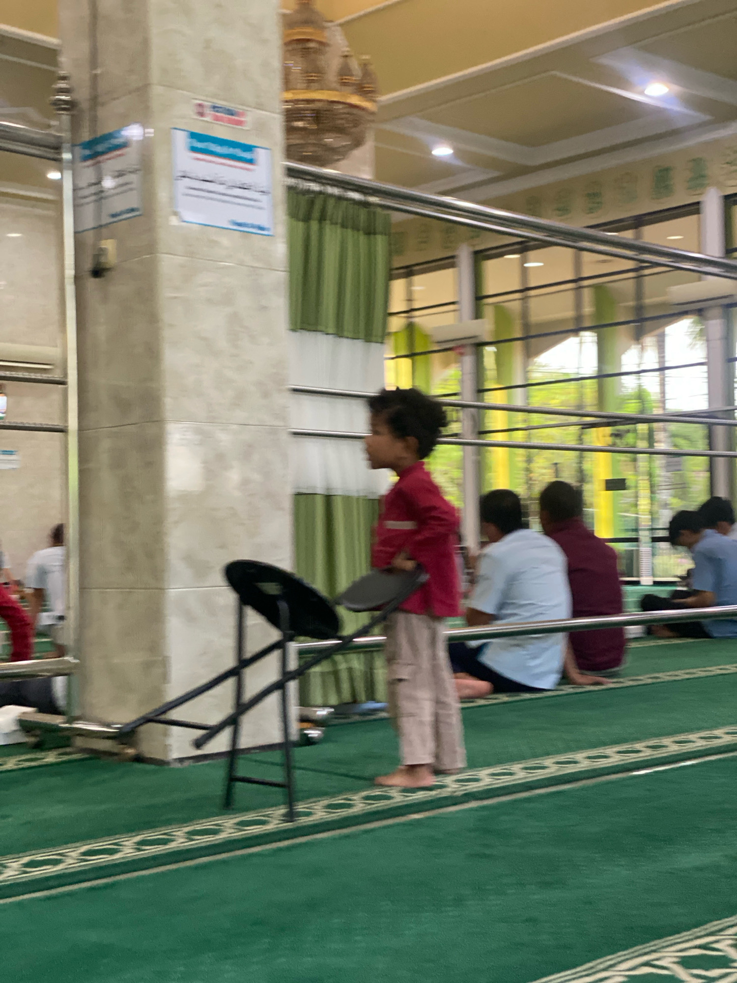The image depicts the interior of a mosque in Banjarbaru, Indonesia, characterized by green carpeting with ornate borders. A young child in a red shirt and light-colored pants stands near a pillar, holding a black folding chair. In the background, several individuals are seated on the floor, facing away from the camera, engaged in prayer or religious observance. The mosque features tall windows allowing natural light to filter in, illuminating the space, and a decorative chandelier hangs from the ceiling. Signs with Arabic script are affixed to the pillar.