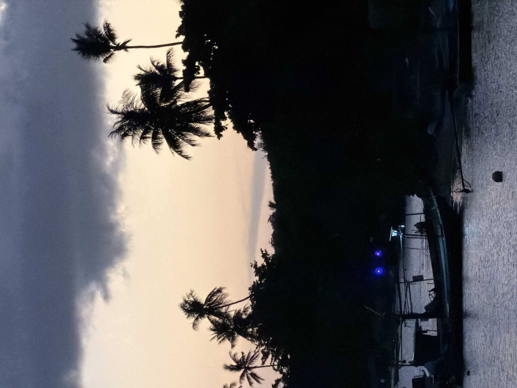 This is a photograph of a harbor in Ko Samui District, Thailand, during twilight. The sky is a gradient of dark gray clouds at the top transitioning to a soft orange and yellow hue near the horizon. Silhouetted against the sky are several palm trees and dense foliage. In the foreground, several boats are docked in the water. One boat in the center has its lights on, emitting a bright blue glow. The water is calm, with subtle ripples reflecting the dim light. The overall scene is dark and atmospheric, suggesting the end of the day.