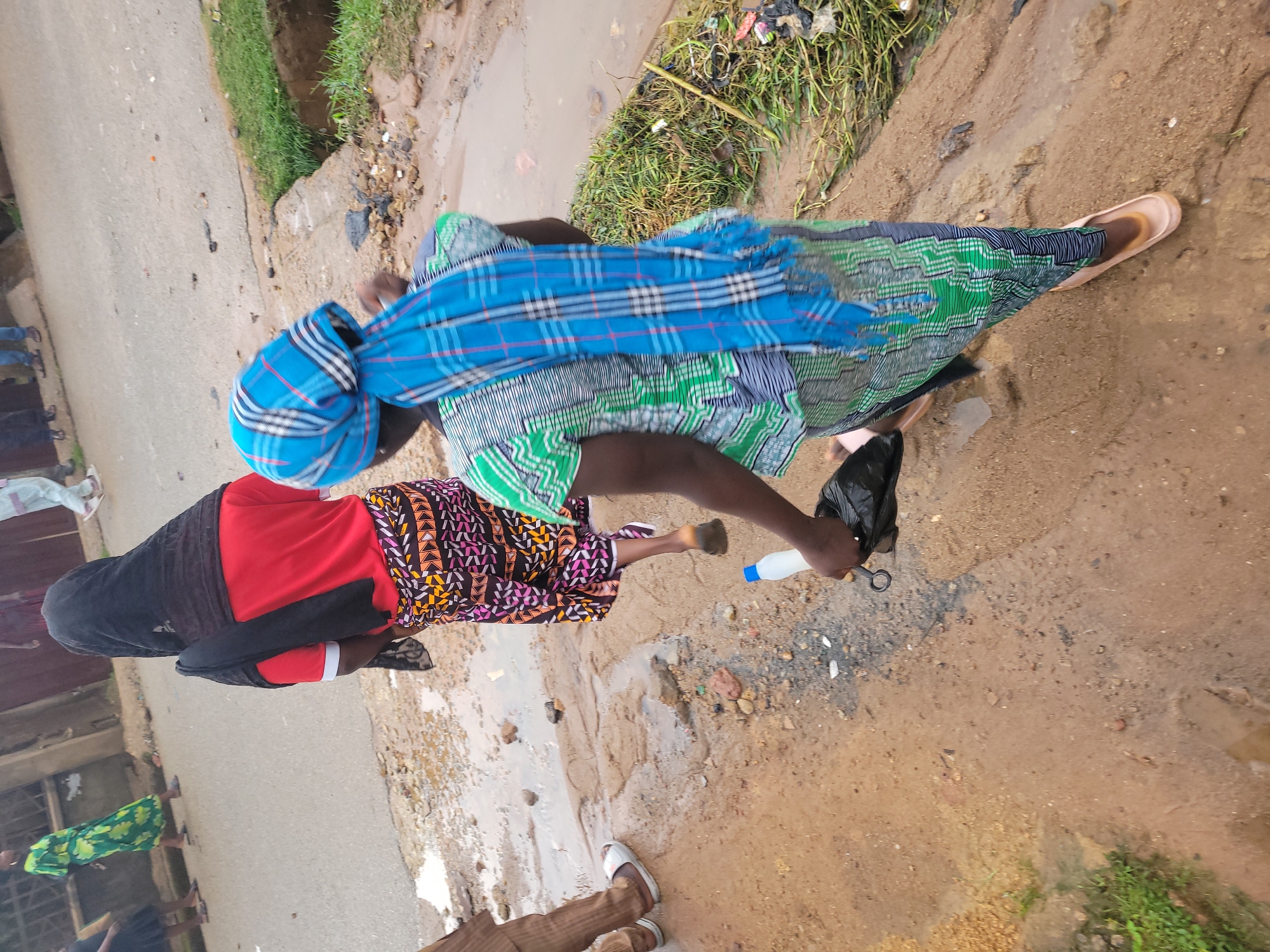 Two women walk on a muddy, uneven path next to a paved road in the city of Jos, Nigeria. The woman in the foreground wears a green and white patterned dress and a blue checkered headscarf. She carries a black plastic bag in her right hand and a white plastic bottle in her left. Behind her, another woman walks wearing a red t-shirt and a black headscarf. She also wears a colorful patterned skirt. The ground is wet and appears to have puddles. The environment suggests recent rainfall or a poorly drained area. Other individuals are visible in the background on the road, some wearing sandals and dresses. The scene depicts a common street view in a developing urban area.