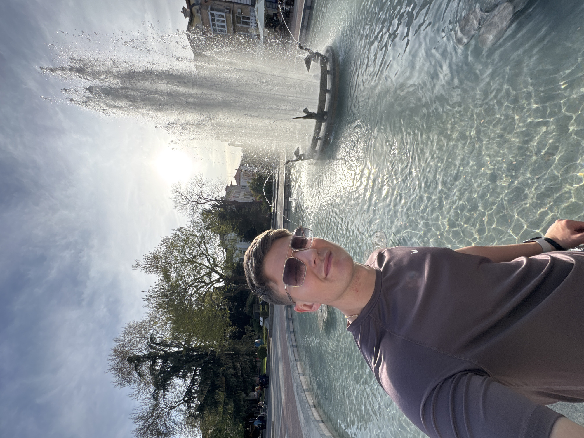 A man in sunglasses and a brown t-shirt stands in front of the Singing Fountains, a landmark in Plovdiv, Bulgaria. He appears to be taking a selfie. The fountain is active, with water spraying upwards. Trees and buildings are visible in the background. The sky is partly cloudy, with sunlight visible through the fountain's water.