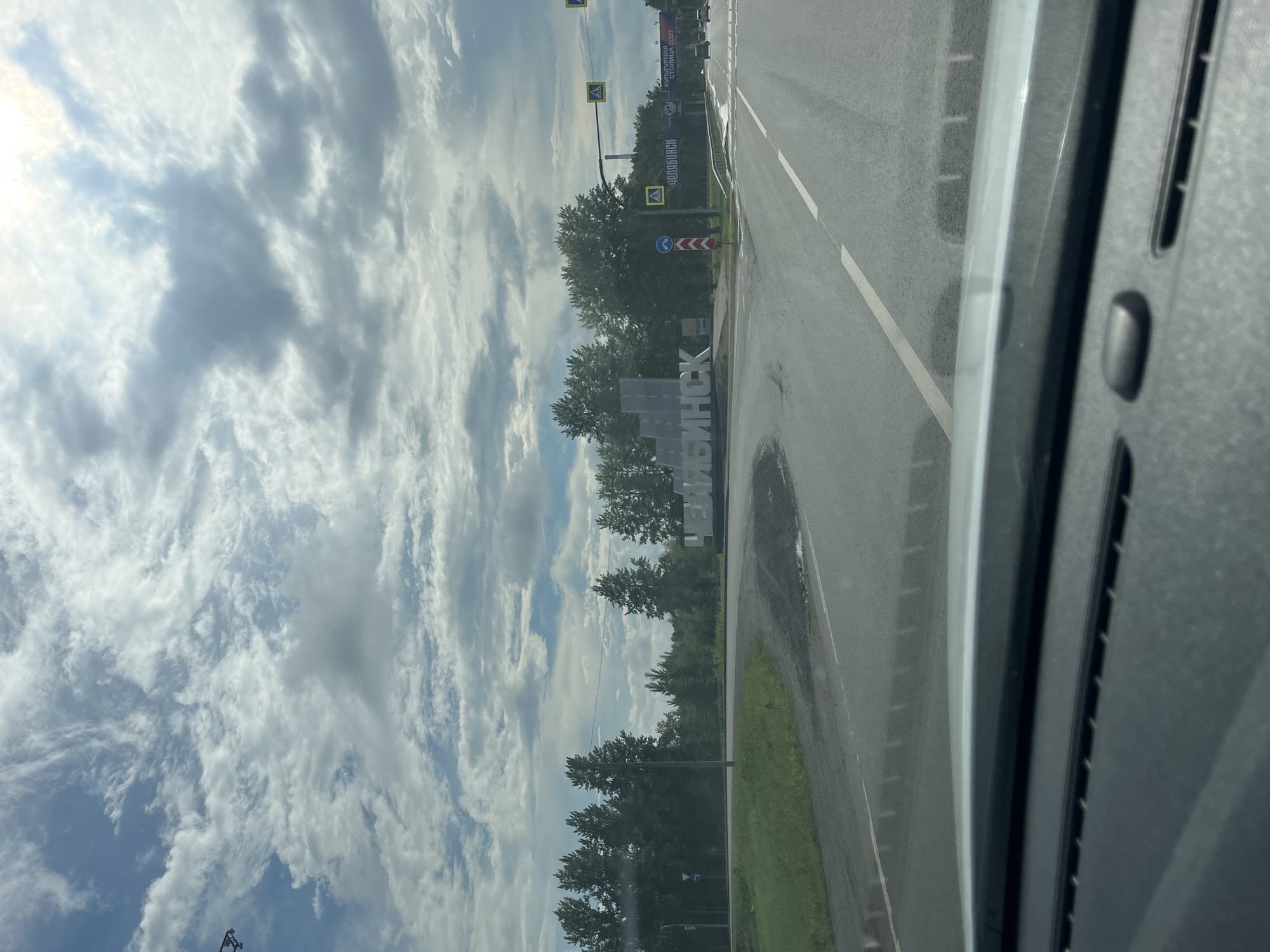 The image shows a road intersection with a large sculpture spelling out "ЧЕЛЯБИНСК" (Chelyabinsk). The sculpture is white and appears to be made of metal or concrete. It is situated in the center of the intersection, surrounded by greenery and trees. The sky is cloudy, with patches of blue showing through. There are road signs and traffic lights visible, indicating this is a public road. The image appears to have been taken from inside a vehicle, looking out through the windshield. The location is identified as Krugloye, Russia, though the name "Chelyabinsk" on the sculpture suggests the scene is on the outskirts of or approaching Chelyabinsk. There are no people visible in the image.