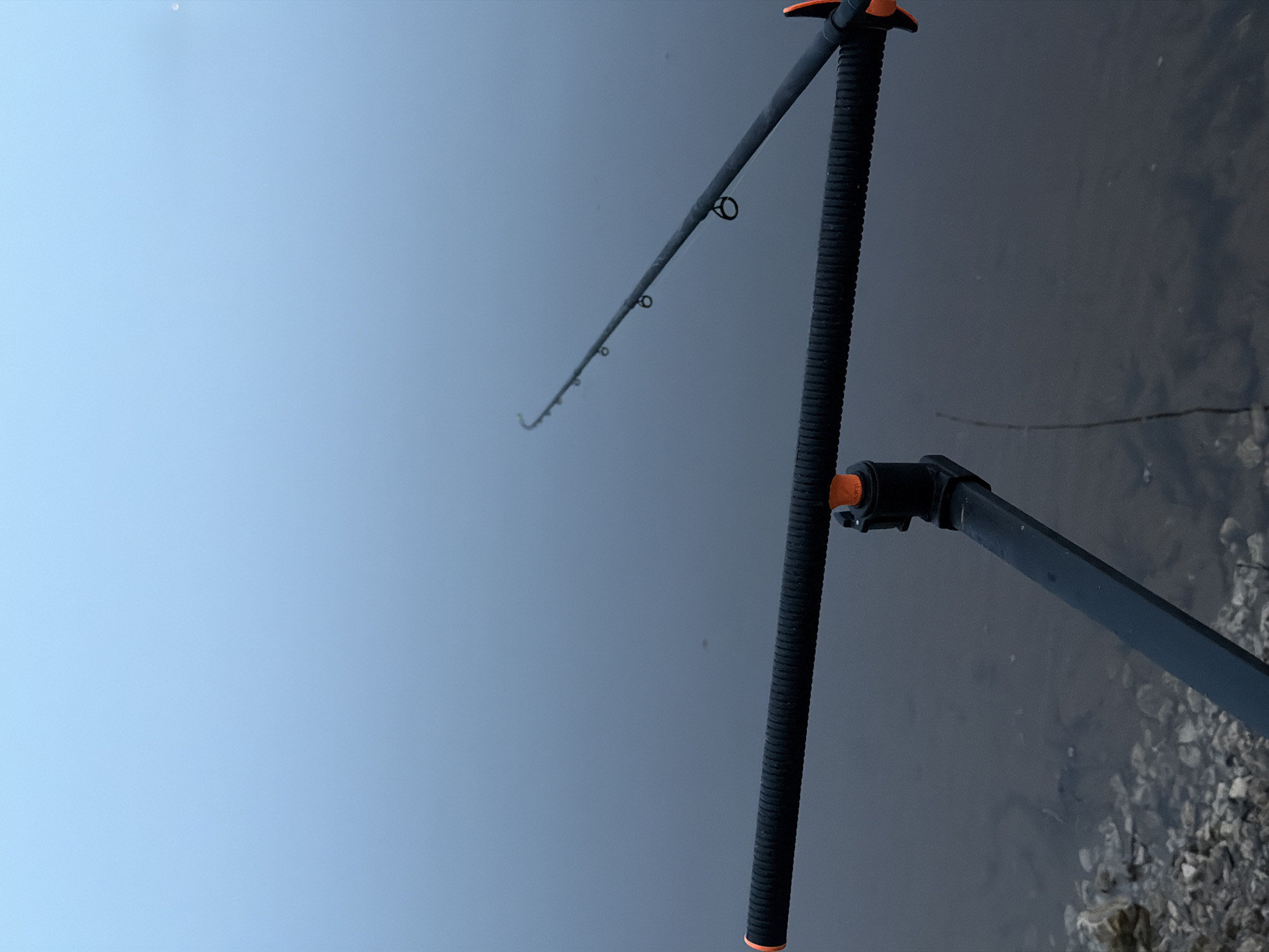 The image, captured in a portrait orientation, features a fishing rod and stand positioned beside a body of water. A black fishing rod with distinct orange accents on its grip and near the reel seat (though the reel itself isn't fully visible) extends upwards and to the left from a black fishing stand. Multiple dark fishing rod guides are visible along the rod's length as it tapers into the distance.
The upper left portion of the image is dominated by a soft, light blue-grey expanse, which could be the sky on an overcast day, thick fog, or a reflection of the sky on an extremely calm water surface. The fishing rod points into this ethereal area. The lower right section of the image reveals the water's edge, where clear water allows visibility of small, light-colored pebbles and rocks on the bottom, indicating a shallow area. The water beyond this immediate shore appears darker, reflecting the muted light conditions.
No people are visible in the scene, and no text is present. The overall atmosphere suggests a tranquil setting, possibly early morning or late evening, by a lake or river near Chaikovsky, Russia, characterized by calm water and subdued lighting.