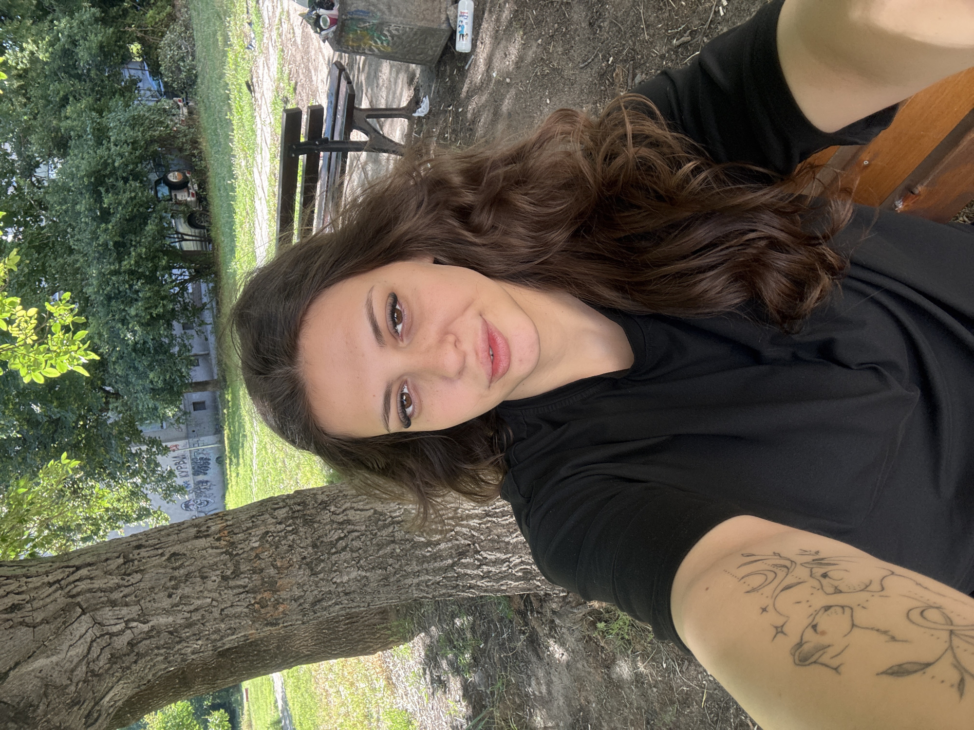 In Plovdiv, Bulgaria, a young woman with long, wavy brown hair and dramatic winged eyeliner takes a selfie outdoors. She is wearing a black t-shirt and has a tattoo of two lions on her left arm. She is sitting on a wooden park bench under the shade of a large tree. In the background, there is more greenery, a building with graffiti, and a trash can. The lighting suggests it is daytime.