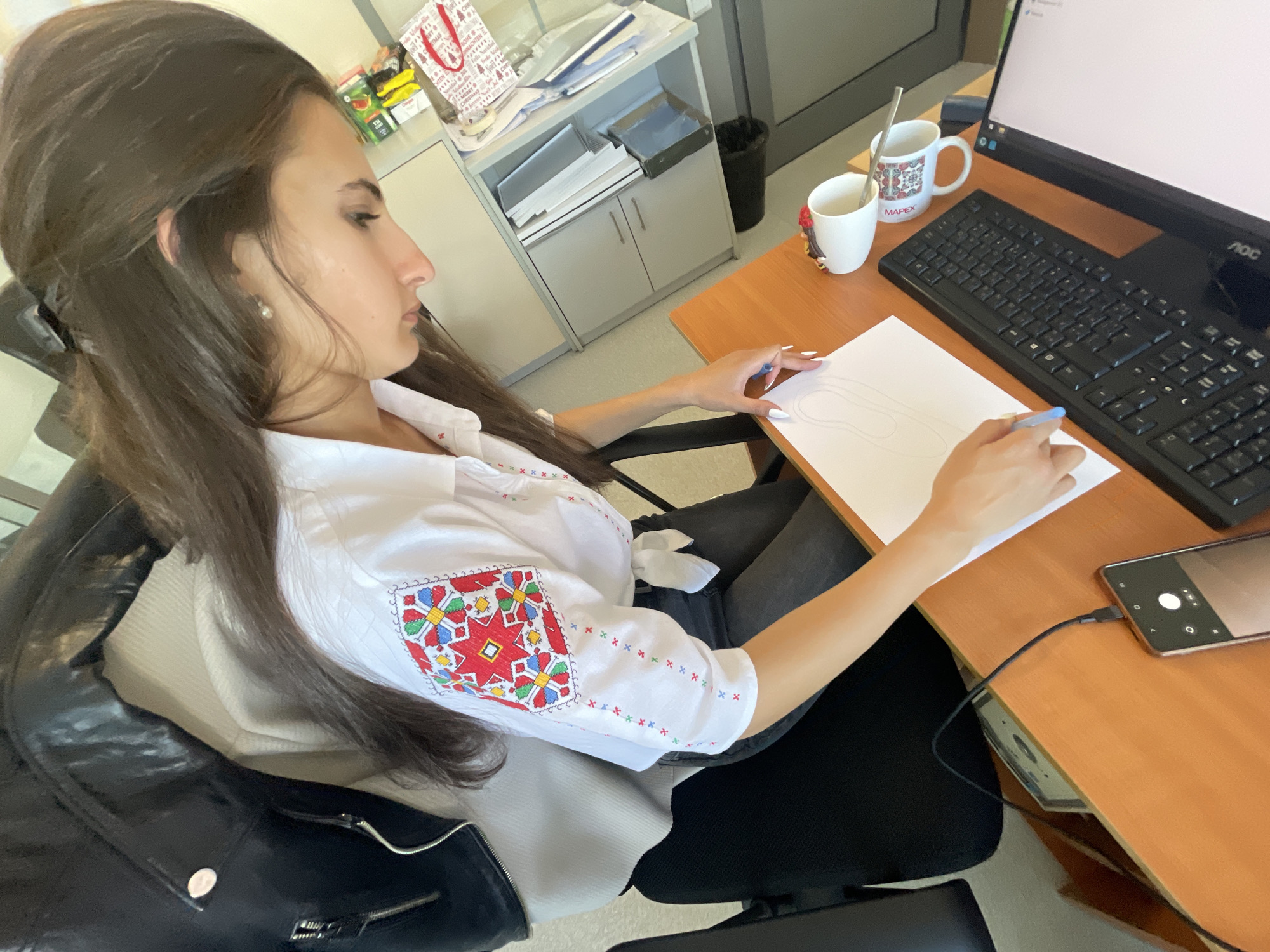 A young woman is depicted in a side profile, seated at a wooden office desk in what appears to be a brightly lit office environment. She has long, dark hair and is wearing a white long-sleeved shirt adorned with vibrant, traditional embroidered patterns in red, blue, green, and yellow on the sleeves and shoulders, along with dark trousers. An earring is visible in her left ear.
Her right hand holds a light blue pen, sketching an outline, possibly of a shoe or slipper, on a white sheet of paper resting on the desk. Her left arm is also on the desk.
On the desk, to her right, is a black computer monitor displaying "AOC" branding, and a black keyboard. Two white mugs are present; one contains a metallic spoon, and the other, featuring a colorful patterned design, has the word "MAPEX" visible on it. A black smartphone, with its screen showing a camera interface, is connected by a charging cable and rests on the desk near the keyboard.
In the background, there's a light-colored office cabinet filled with documents and files, and a black waste bin. A red and white gift bag with "MERRY CHRISTMAS" text is placed on top of the cabinet, alongside some small packets or boxes. The overall scene suggests a focused work or creative activity taking place during the daytime in an office setting.