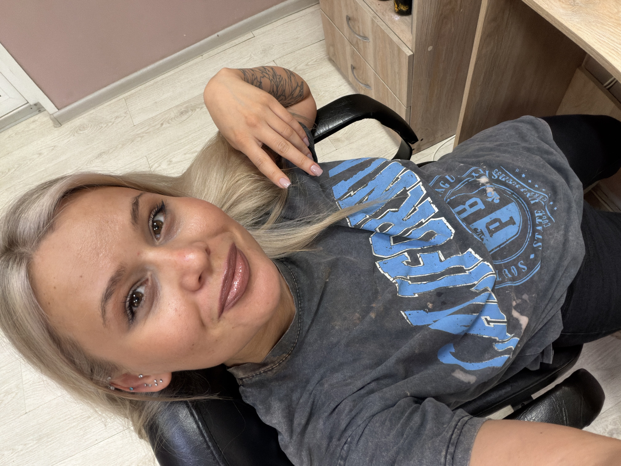 A woman with blond hair is sitting in a black office chair. She is wearing a gray t-shirt with a blue graphic print, and black pants. She has a tattoo on her left arm and multiple piercings in her left ear. The woman is looking at the camera and smiling. The background includes a light-colored wooden desk or cabinet, a light pink wall, and a light-colored wooden floor. The photograph appears to have been taken from a high angle. The location is presumed to be in Sarapul, Russia.